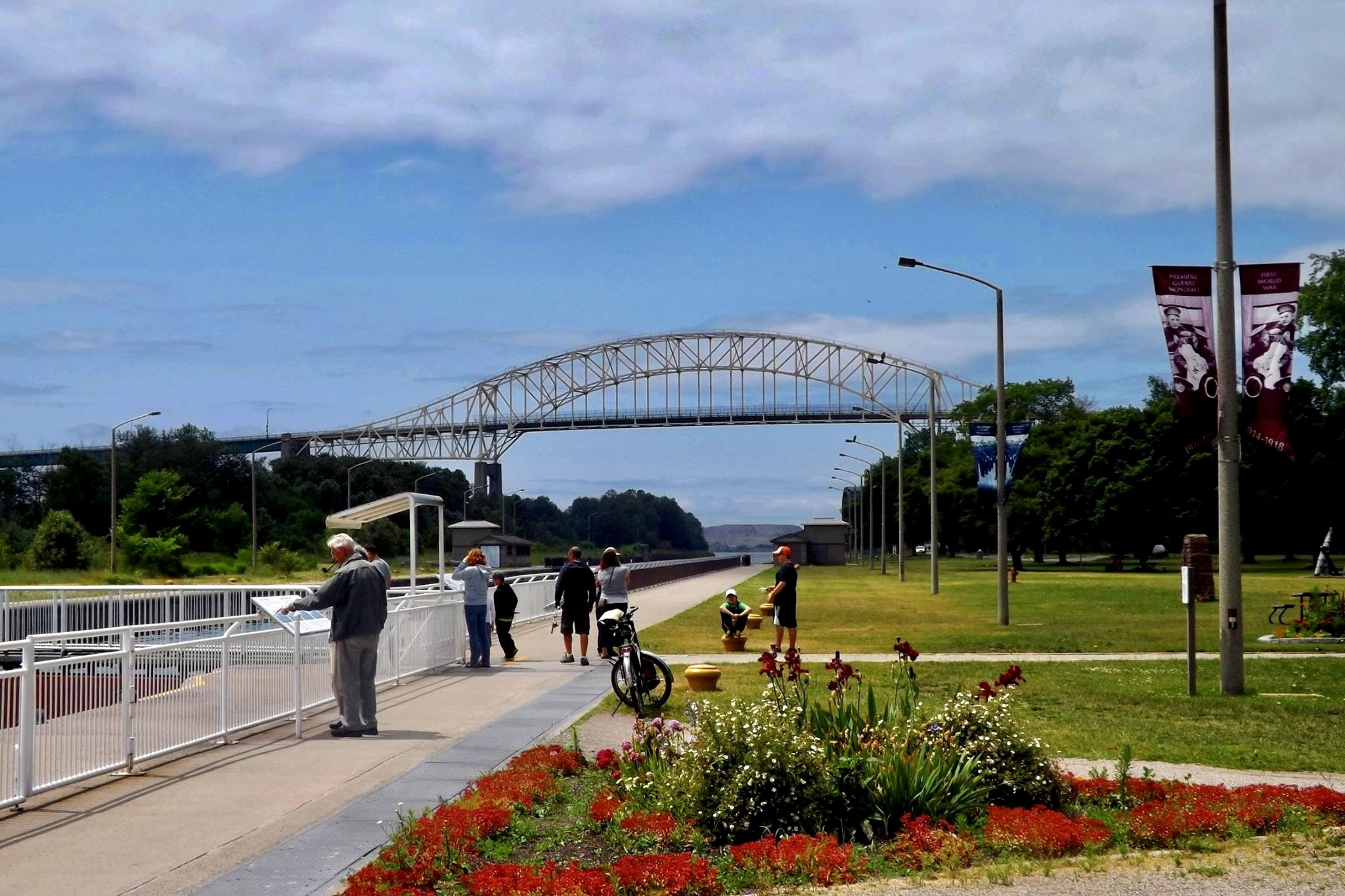 A truss arch bridge spans a river in the background, with a flower-lined pedestrian walkway and lamp posts in the foreground.