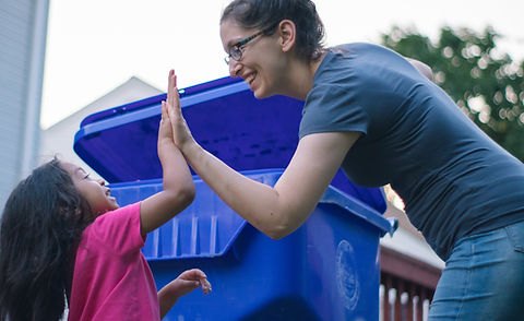 A woman and child high-five in front of a large blue wheeled recycling bin outdoors.