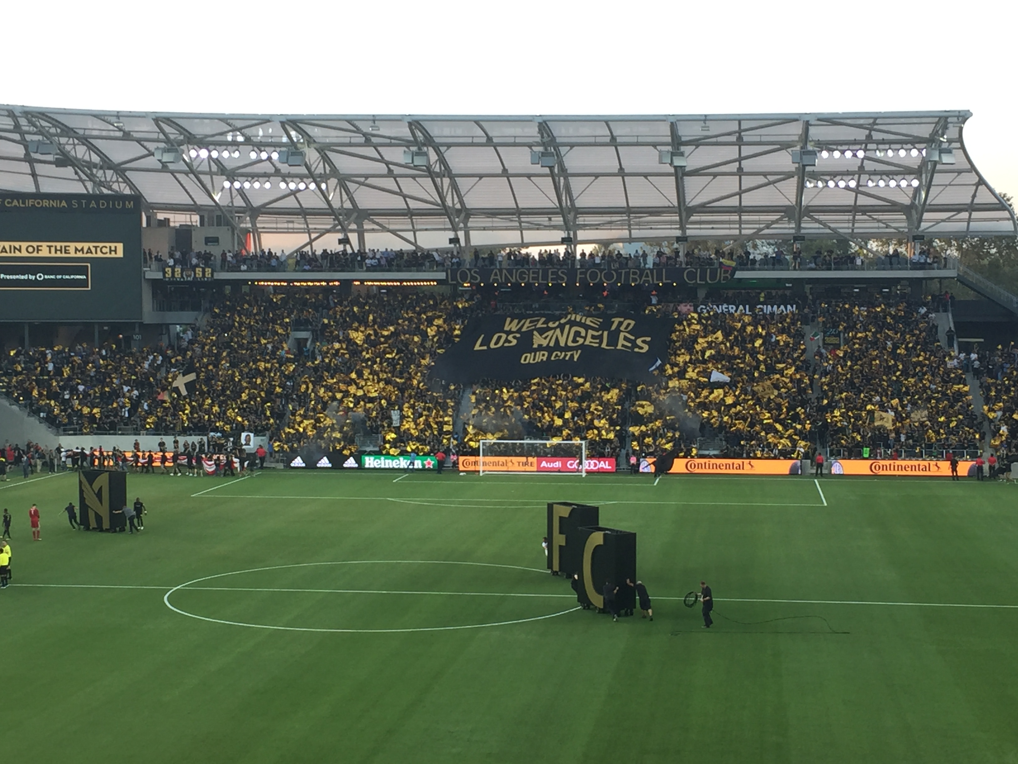 Thousands of fans in a stadium supporter section hold up a massive yellow-and-black tifo banner.