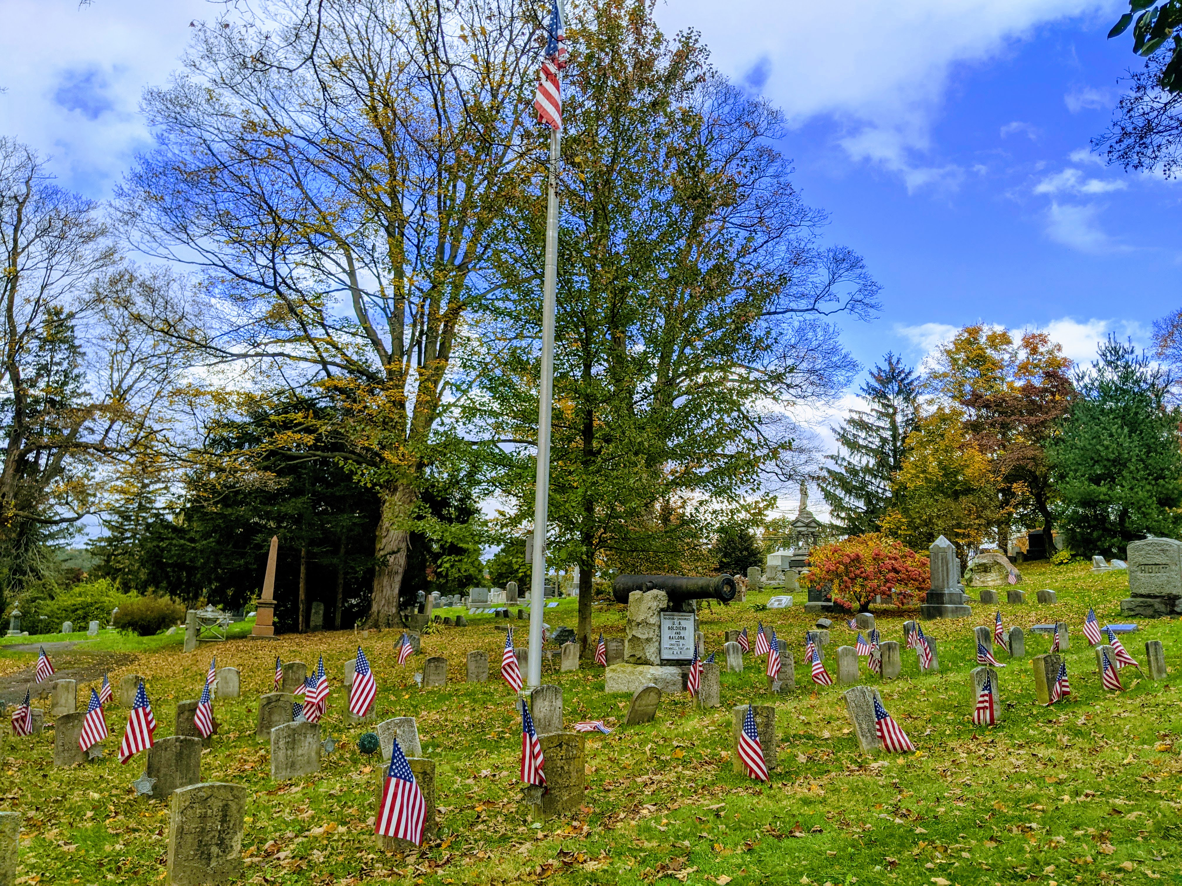 A cemetery with small American flags at grave markers and a flagpole flying a full-size flag, surrounded by autumn trees.