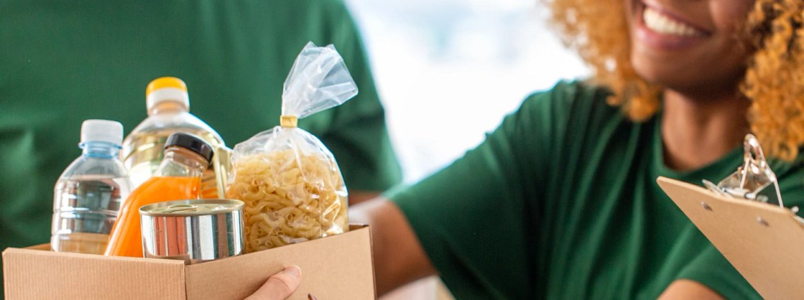 Smiling volunteer in green shirt holds a cardboard box of food donations.