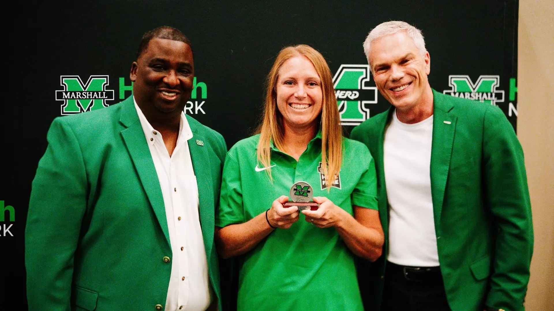 Woman in green polo holds a medallion award, flanked by two men in green blazers before a branded backdrop.