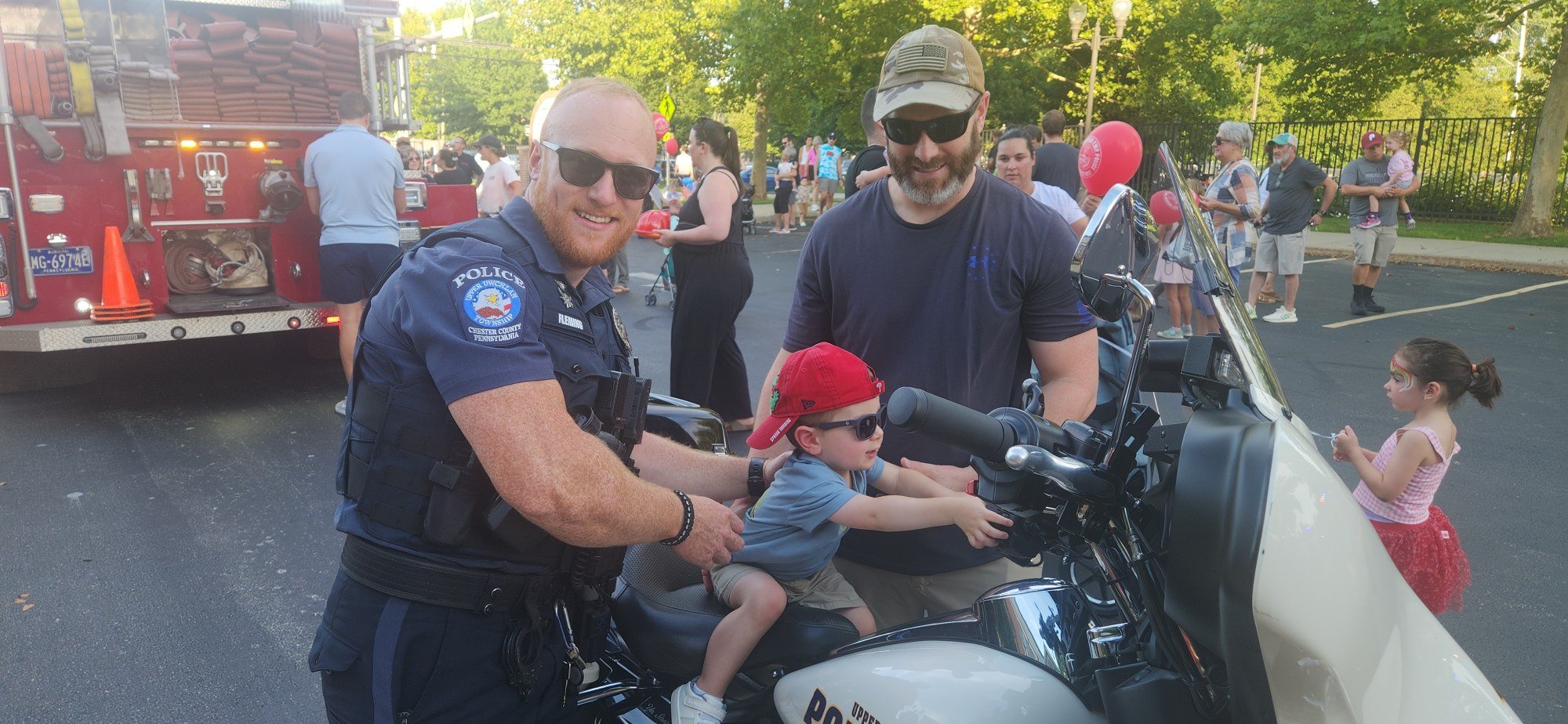 A uniformed officer and a man stand beside a police motorcycle with a toddler seated on it.