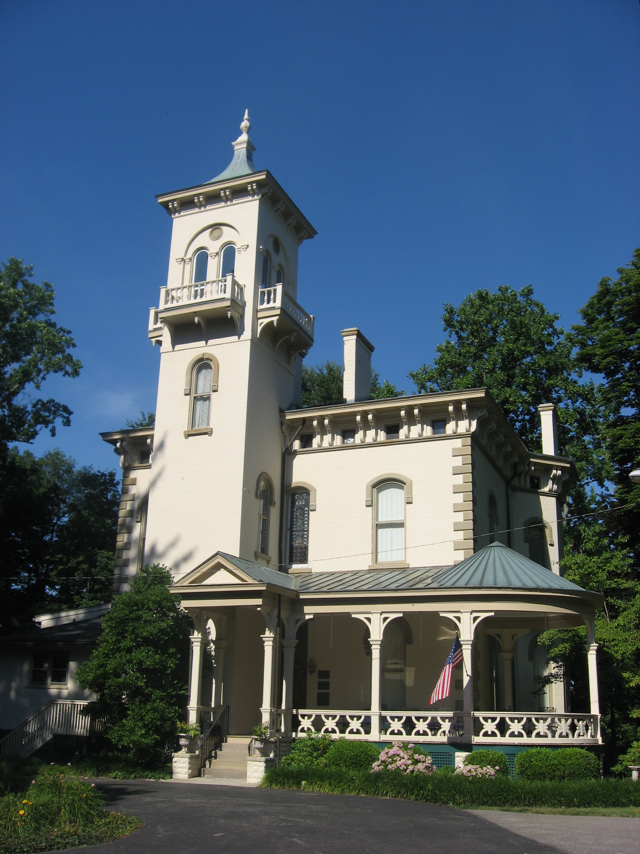Cream-colored Italianate Victorian mansion with tall central tower, wraparound porch, and American flag.