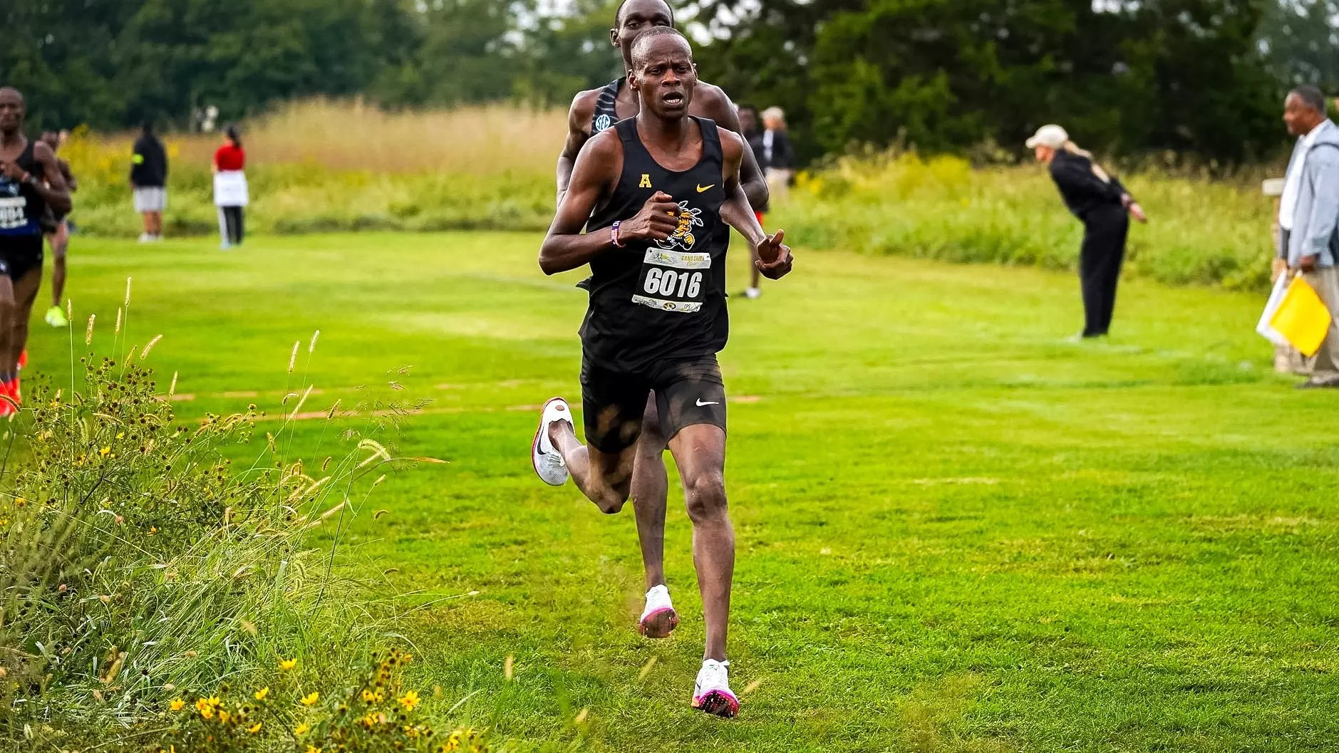A runner in a black singlet leads a cross country race on a grass course.