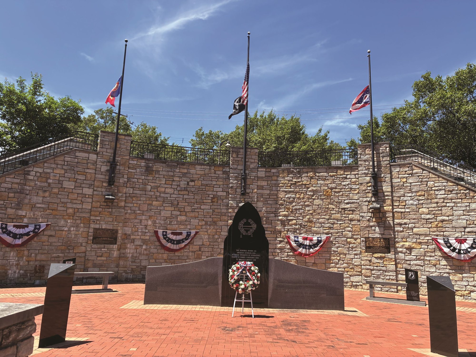 Outdoor veterans memorial plaza with flagpoles, a stone monument, and a floral wreath.