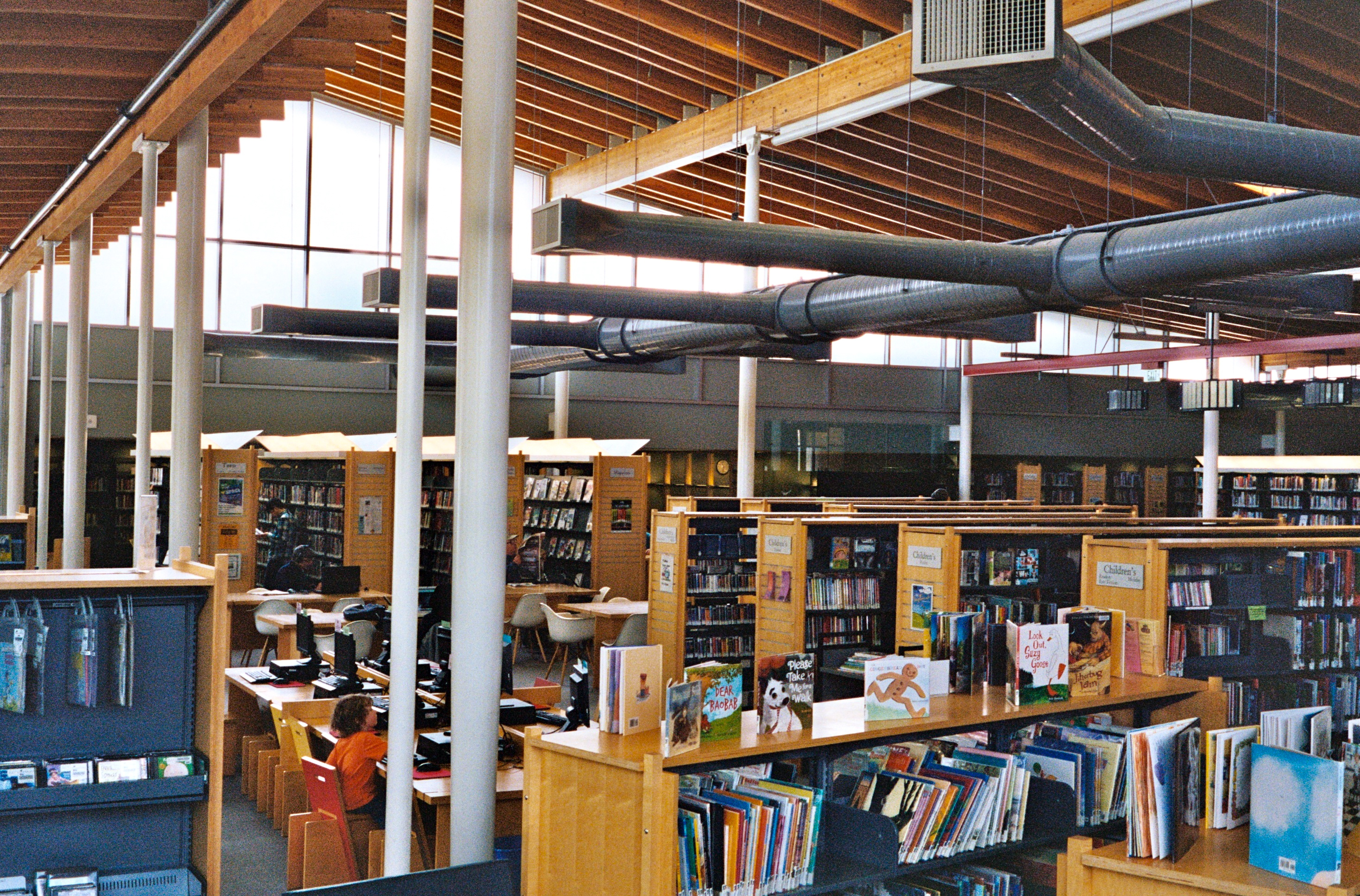 Interior of a public library with bookshelves, reading tables, and high ceilings with exposed ductwork.