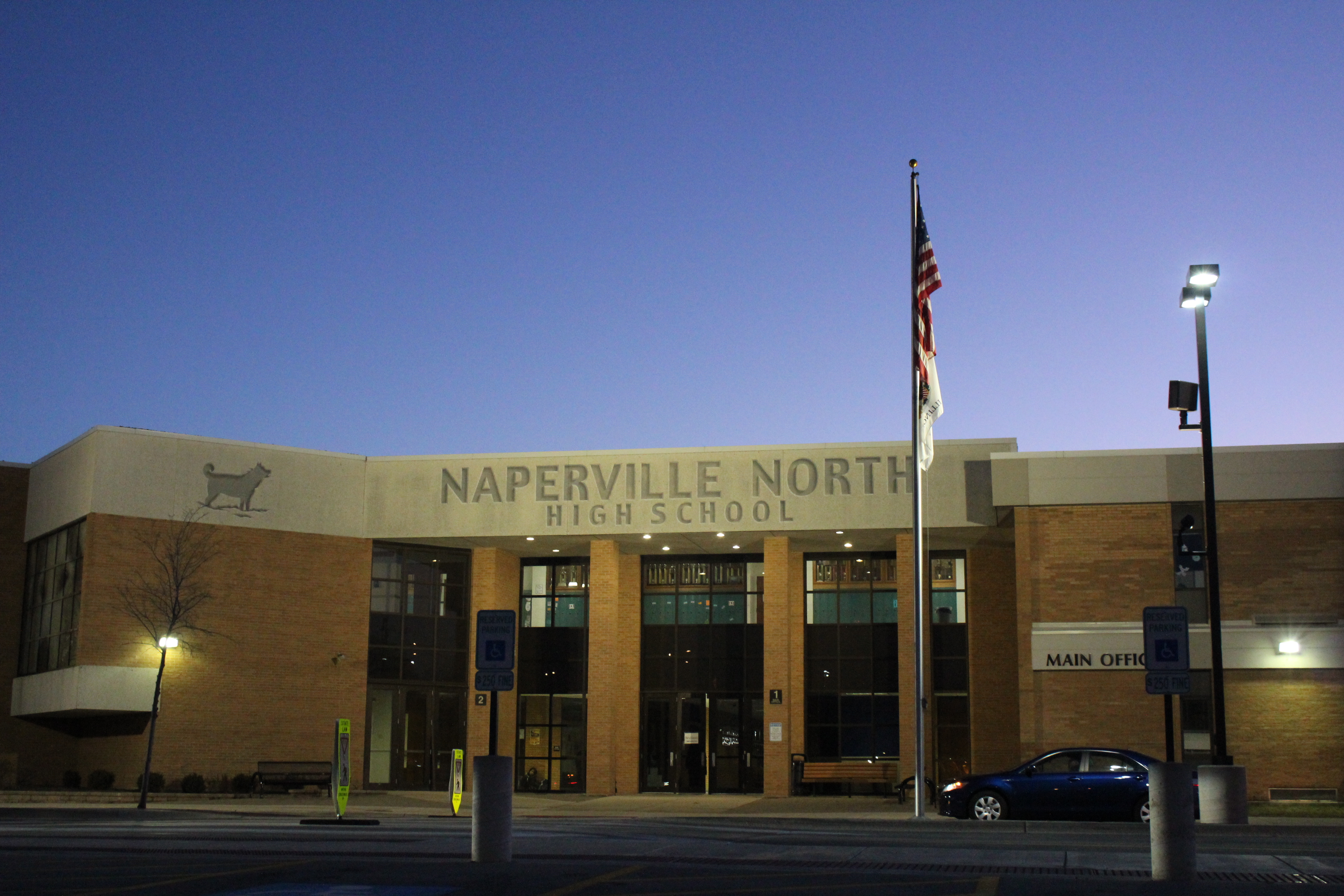 Illuminated main entrance of a brick high school building at dusk with a flagpole.