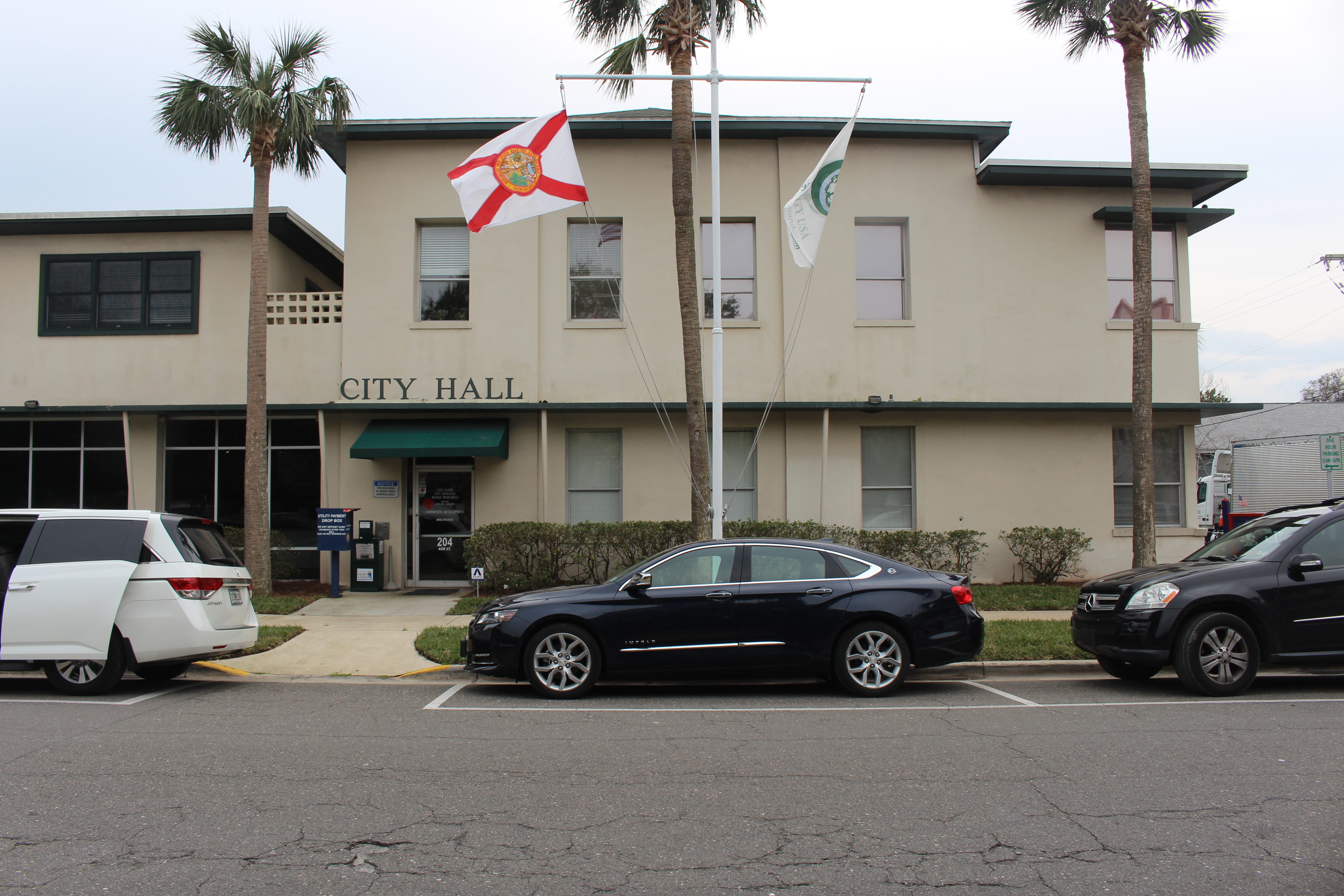 Two-story beige City Hall building with flagpoles and palm trees out front.