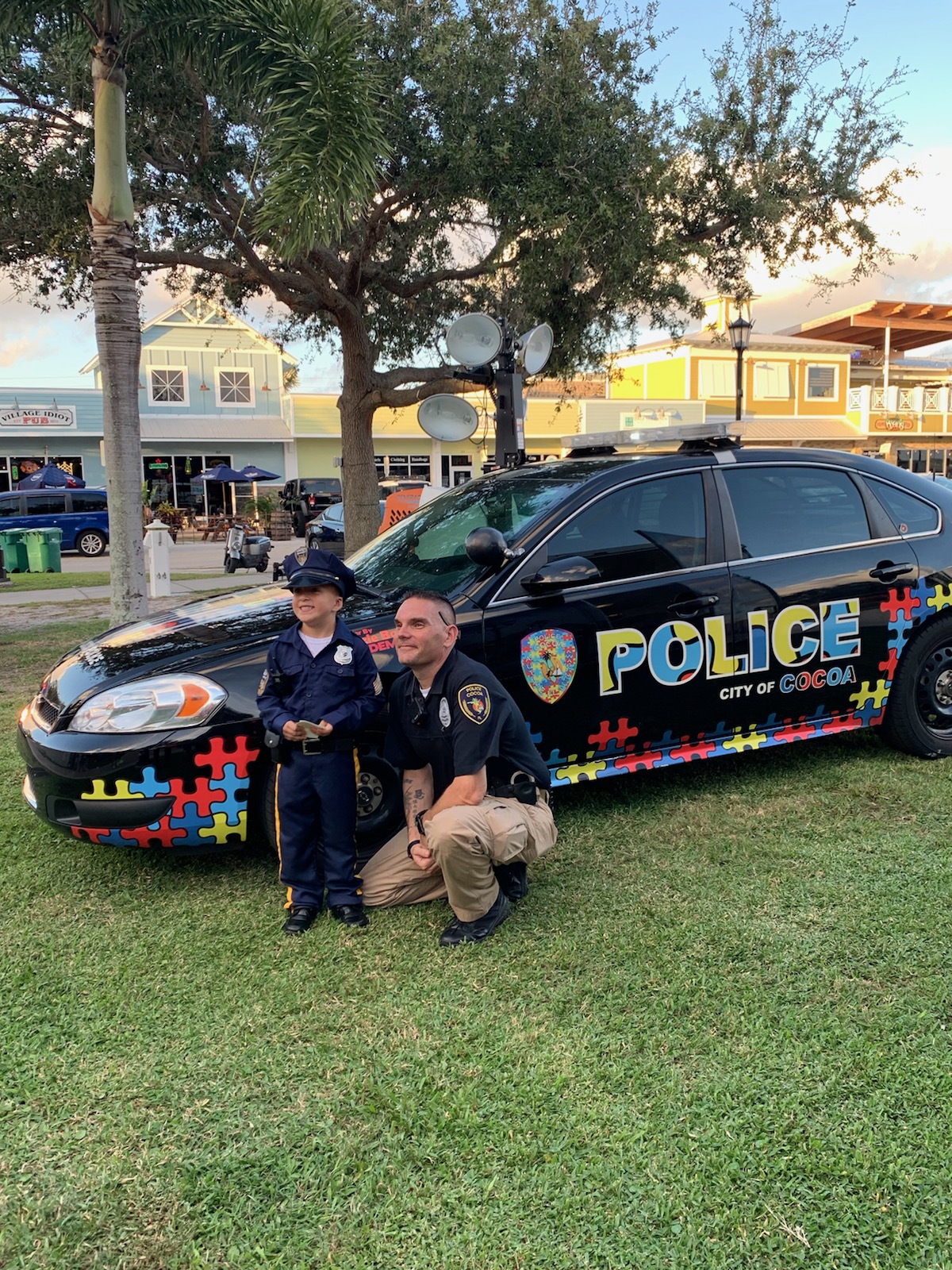 Police officer and child pose beside a patrol car with colorful autism awareness puzzle-piece graphics.