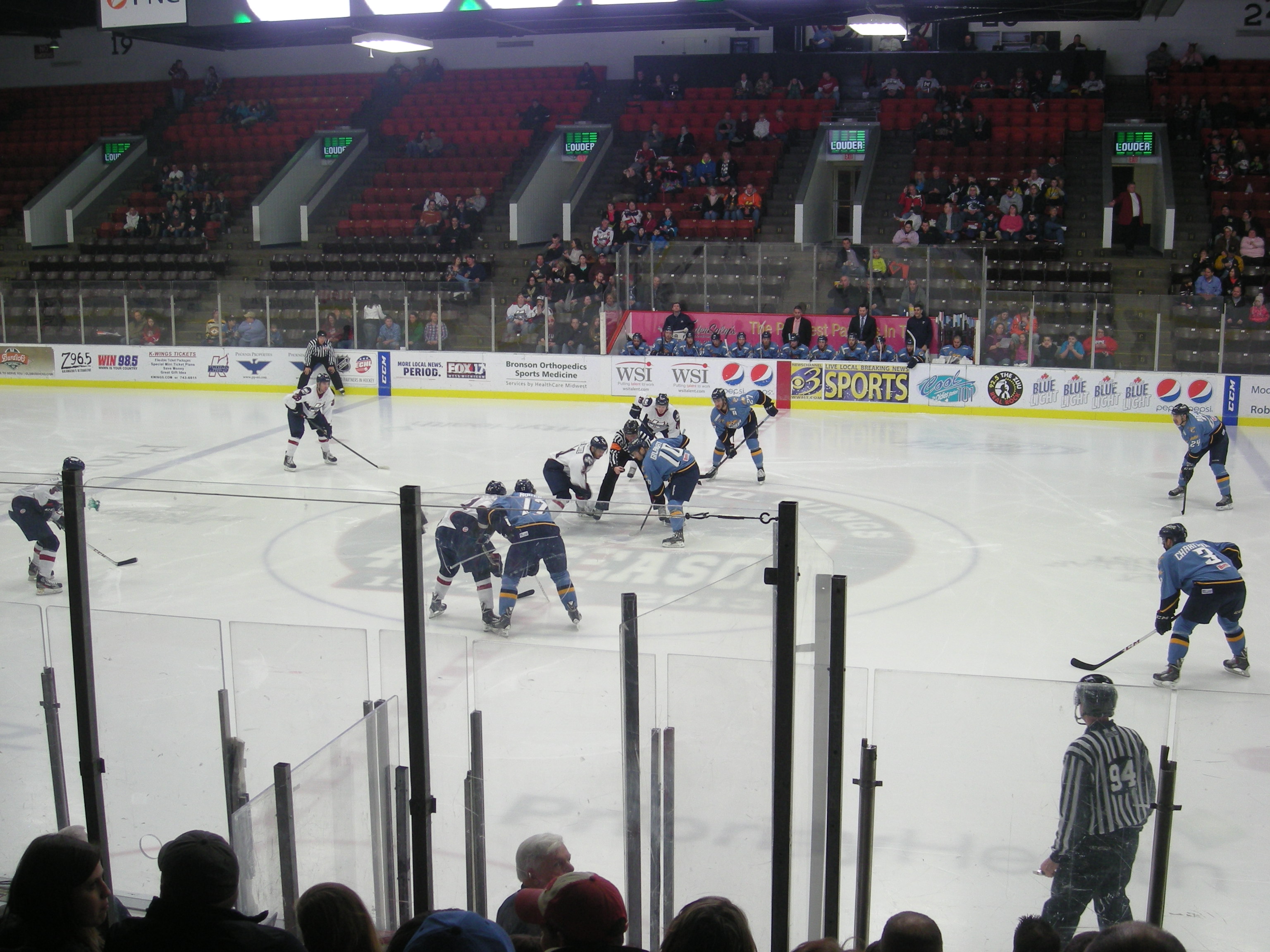 Hockey players in white and dark jerseys compete near center ice at an indoor arena.