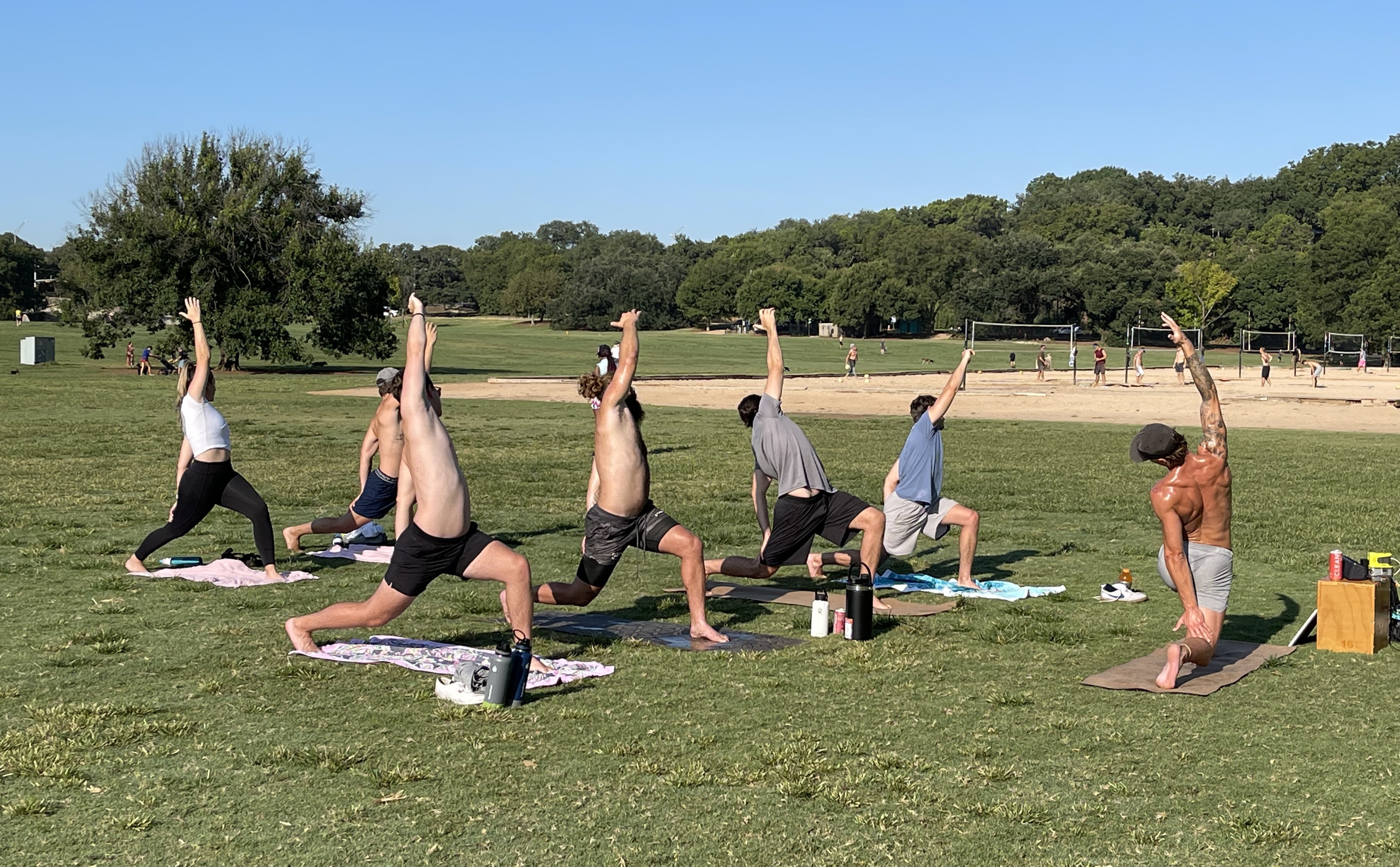 Group of people in a low lunge yoga pose on mats in a sunny park.