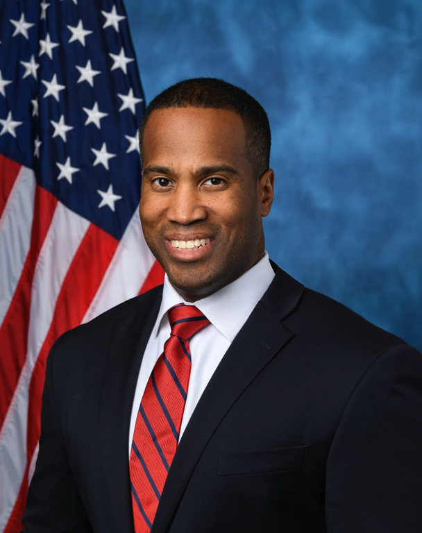John James wearing a dark suit and red striped tie beside an American flag.