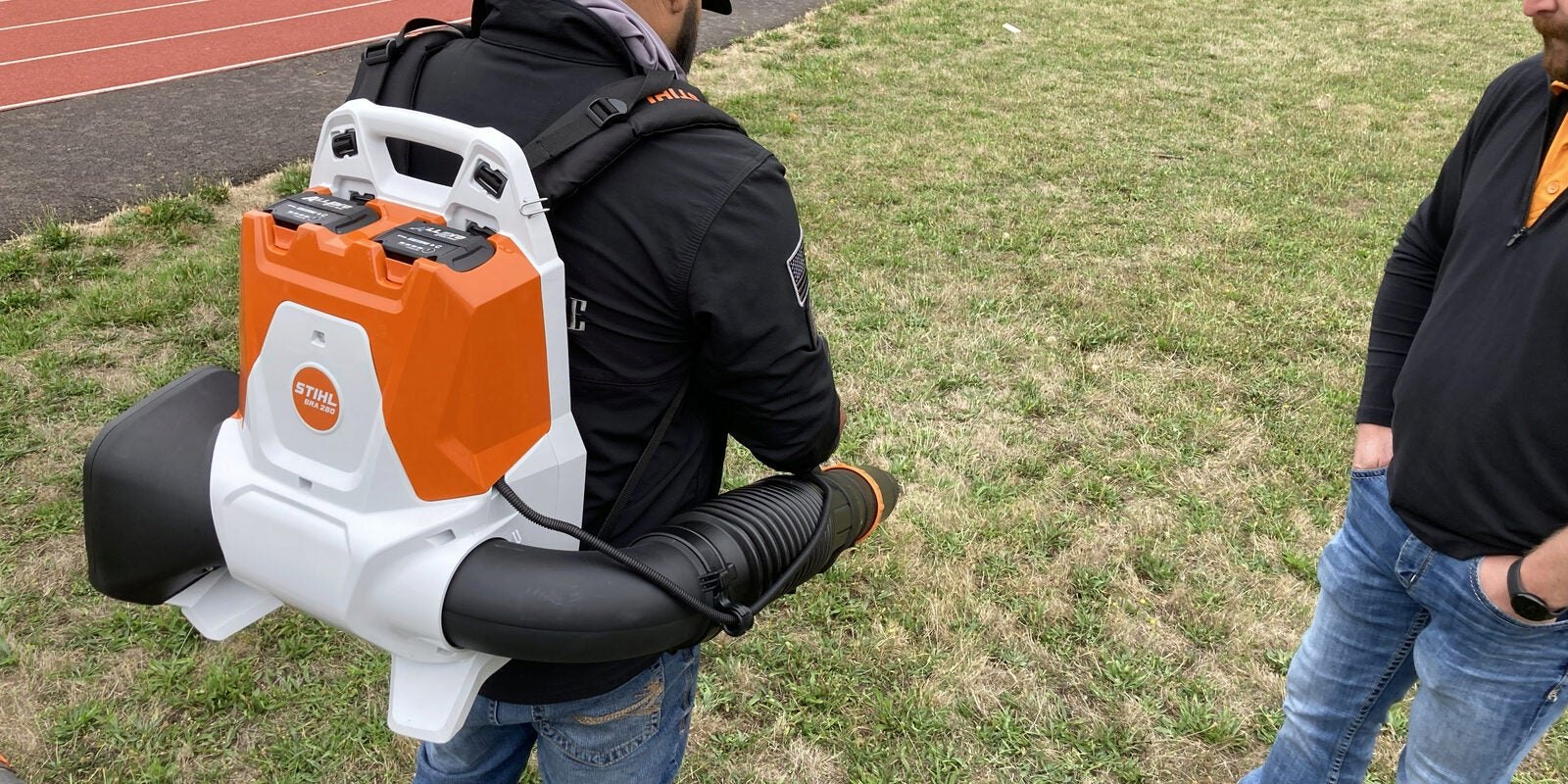 Person wearing an orange-and-white backpack electric leaf blower on a grassy field.