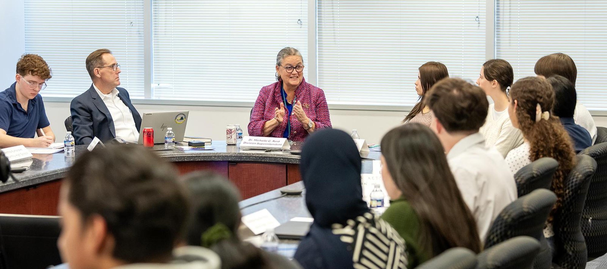 A woman speaks at the head of a curved conference table to a room of seated students.