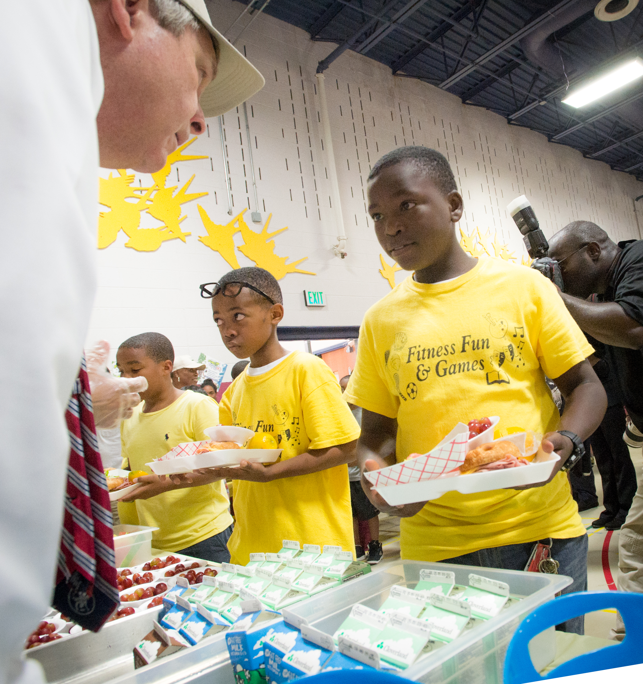 Children in yellow shirts hold food trays in a lunch service line.