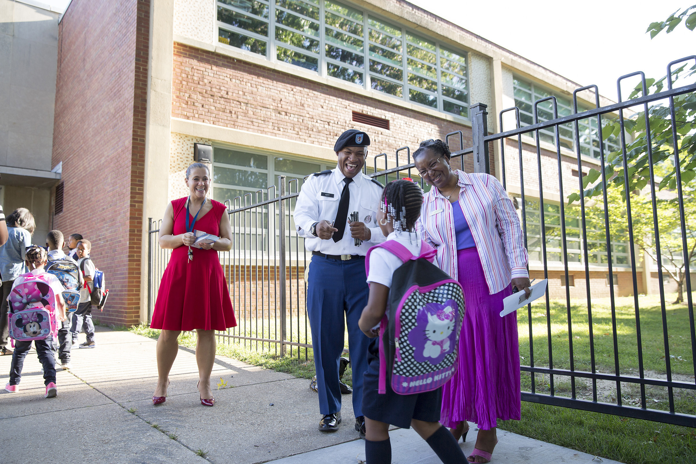 Adults greet young children with backpacks at an elementary school entrance.