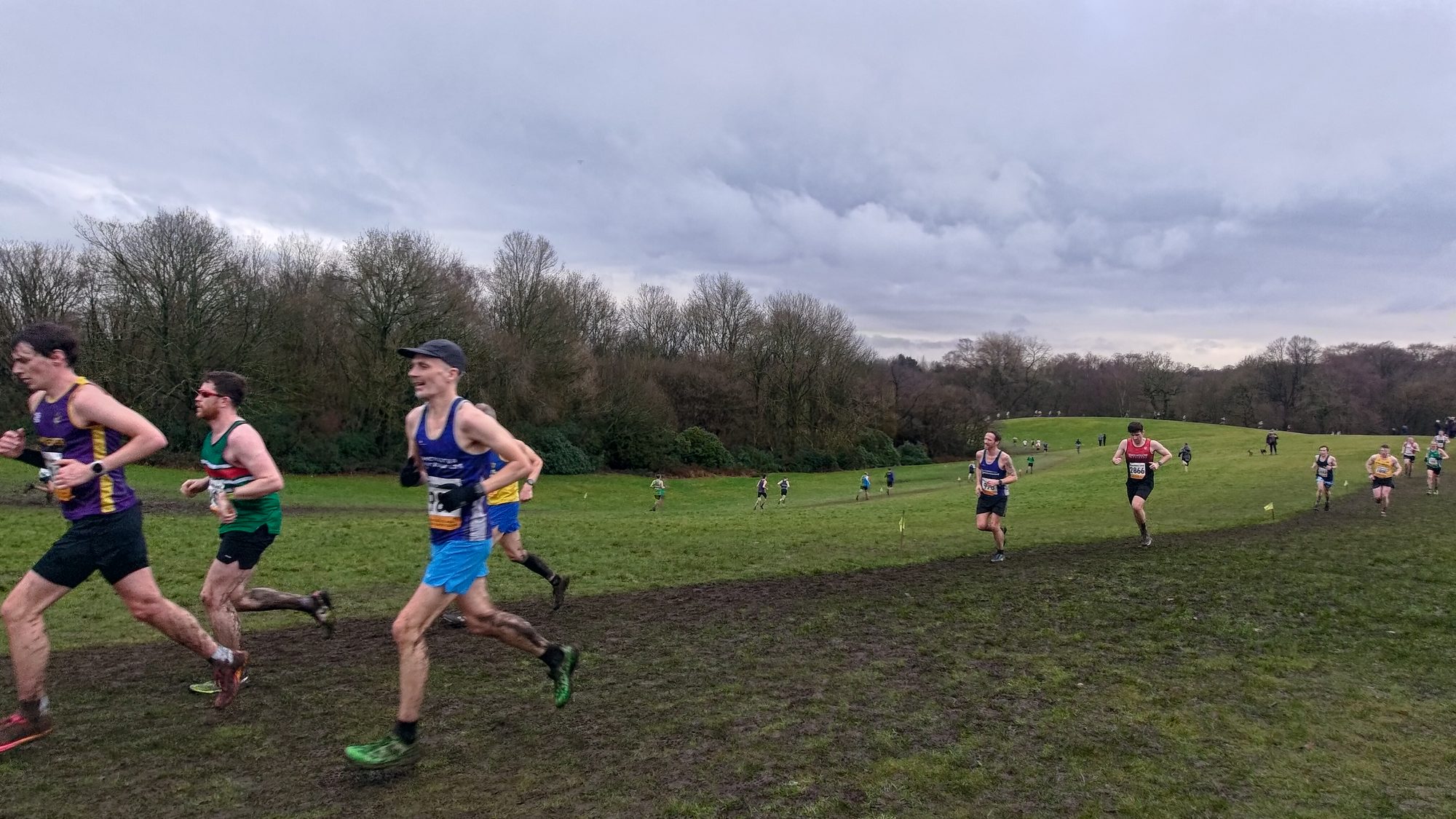 At Manchester Area Cross League on Saturday 7th February 2026, runners tackle the mud of Heaton Park. There is a green yet muddy field, hills visible and trees/greenery. Small yellow flags mark the route. The runners are wearing vests of their club.