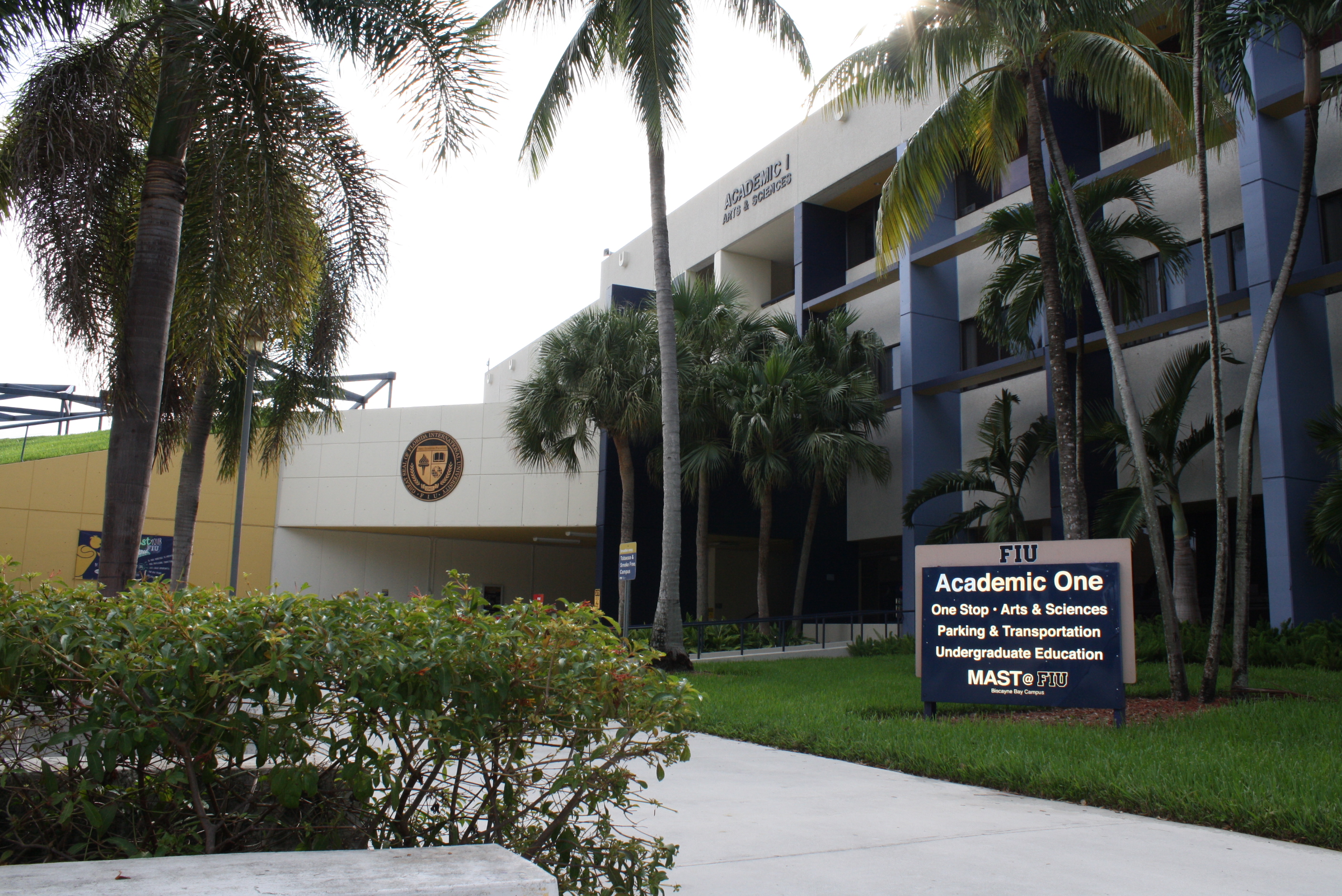 Multi-story university building with palm trees and a sign listing campus departments.