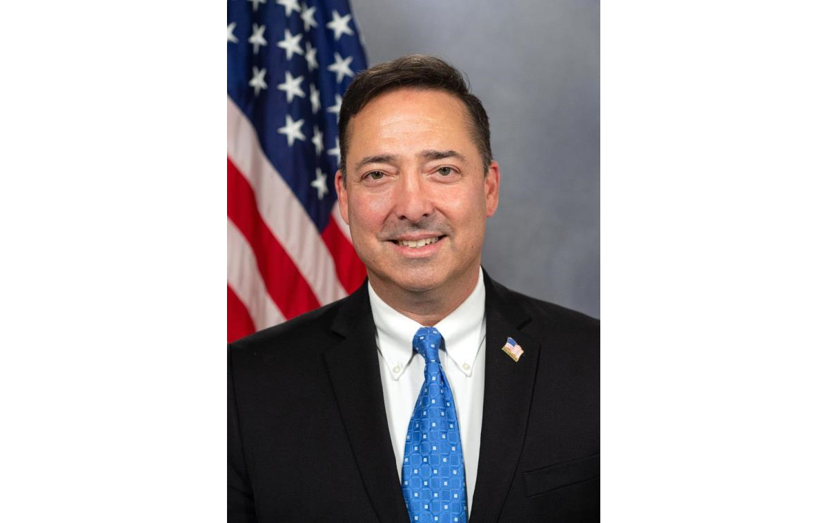Man in a dark suit and blue tie poses for a formal portrait in front of an American flag.