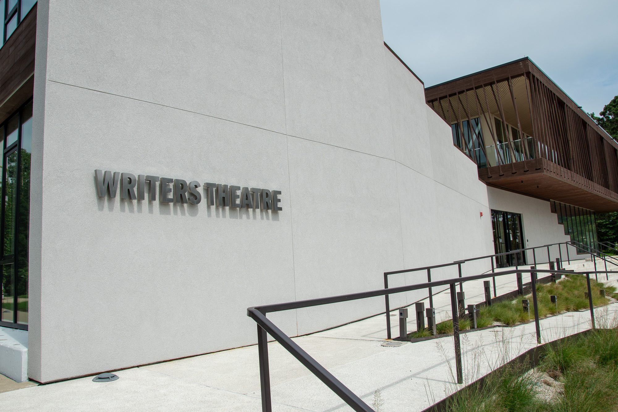Glencoe civic theater. The exterior of Writers Theatre, a modern building with a white stucco facade and dark metal lettering spelling out the venue's name. The structure features warm wood-paneled upper levels, a concrete ramp with metal railings, and landscaped grounds under an overcast sky.