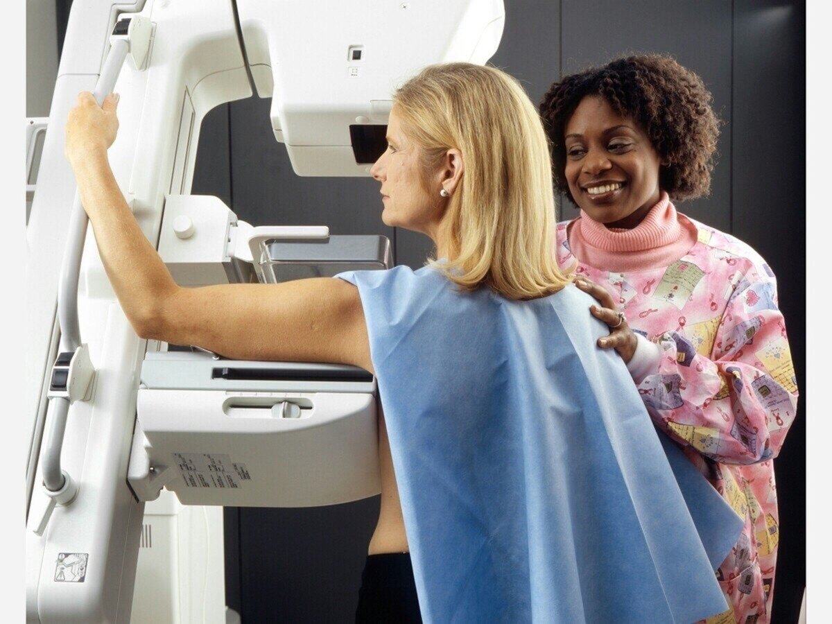 A woman in a patient gown stands at a mammography machine while a technician assists her.