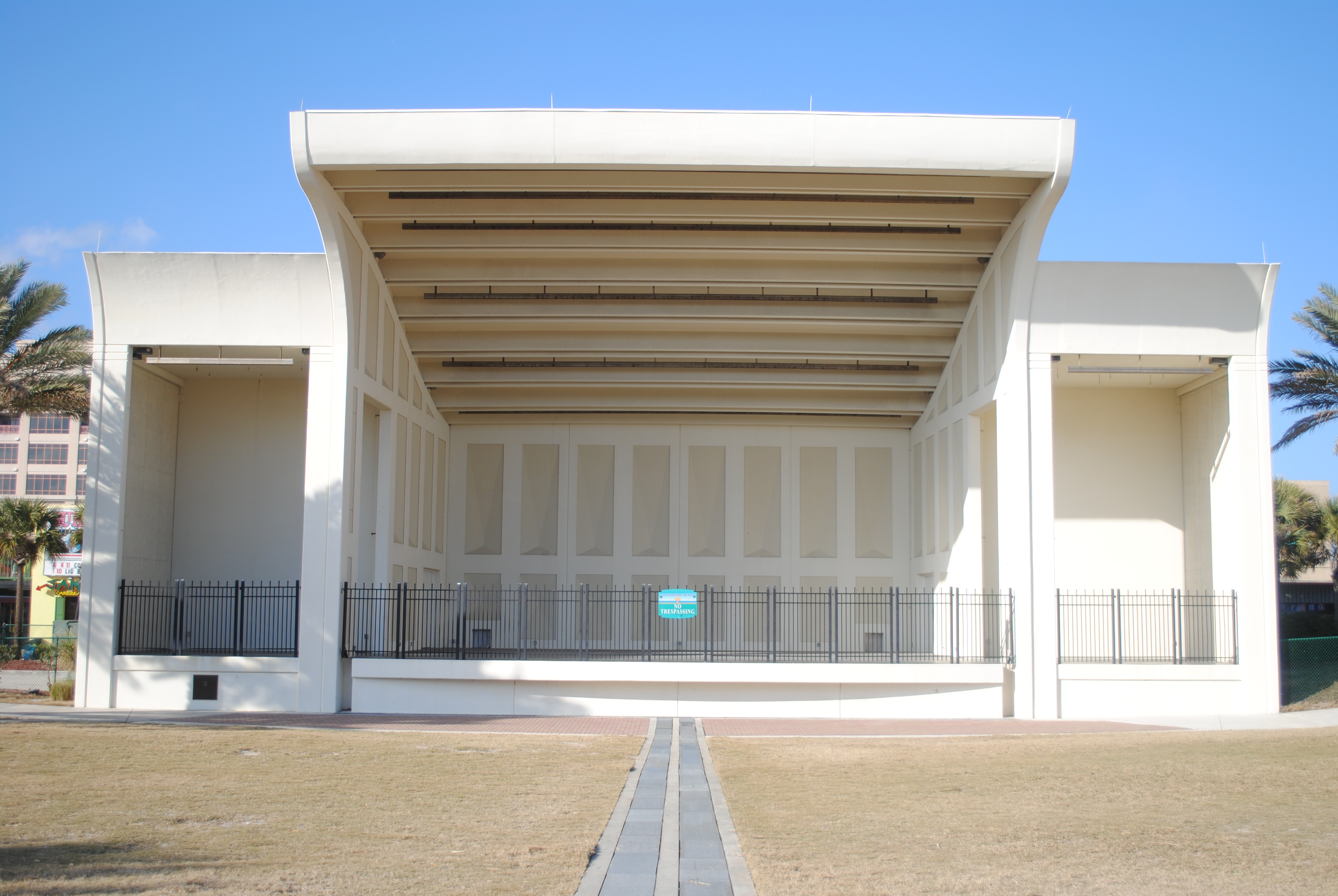 Open-air pavilion with a large cantilevered roof, palm trees, and a brick pathway on a sunny day.