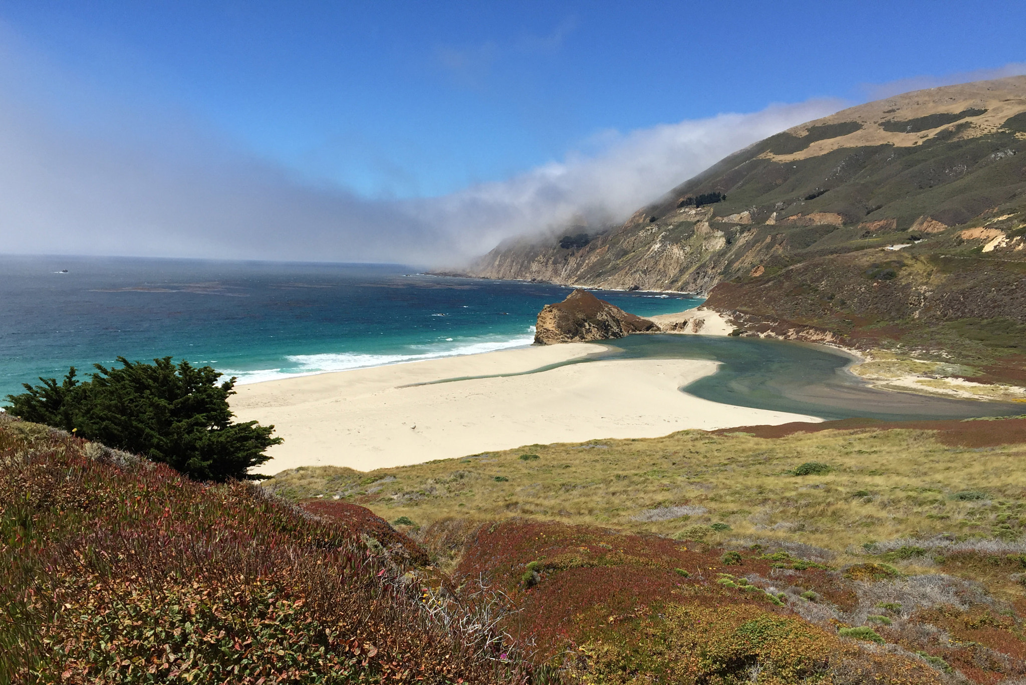 A sweeping coastal view of a sandy beach, turquoise ocean, and fog-covered green hillsides.