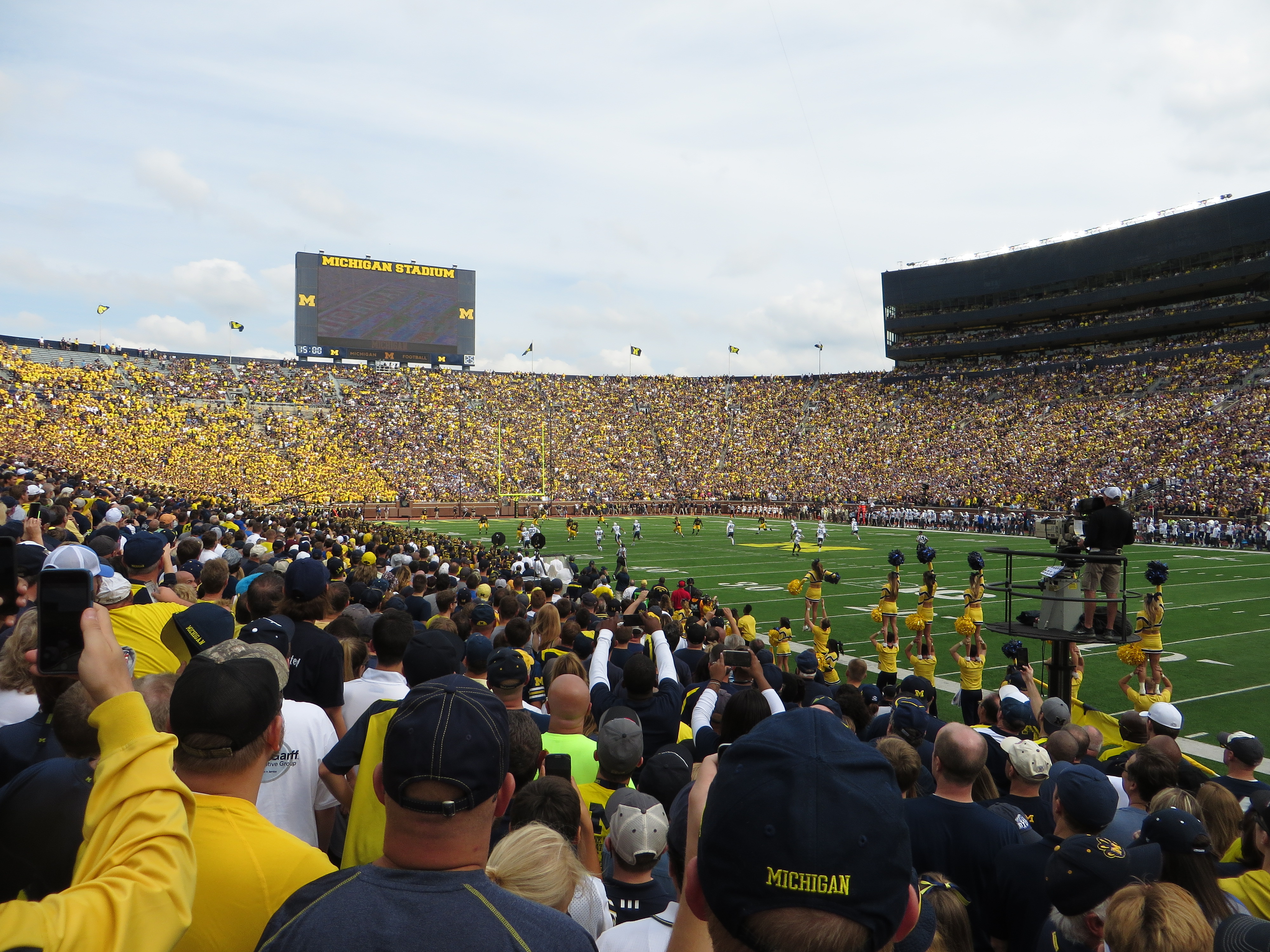 Packed football stadium with fans in yellow and blue, scoreboard reading Michigan Stadium.