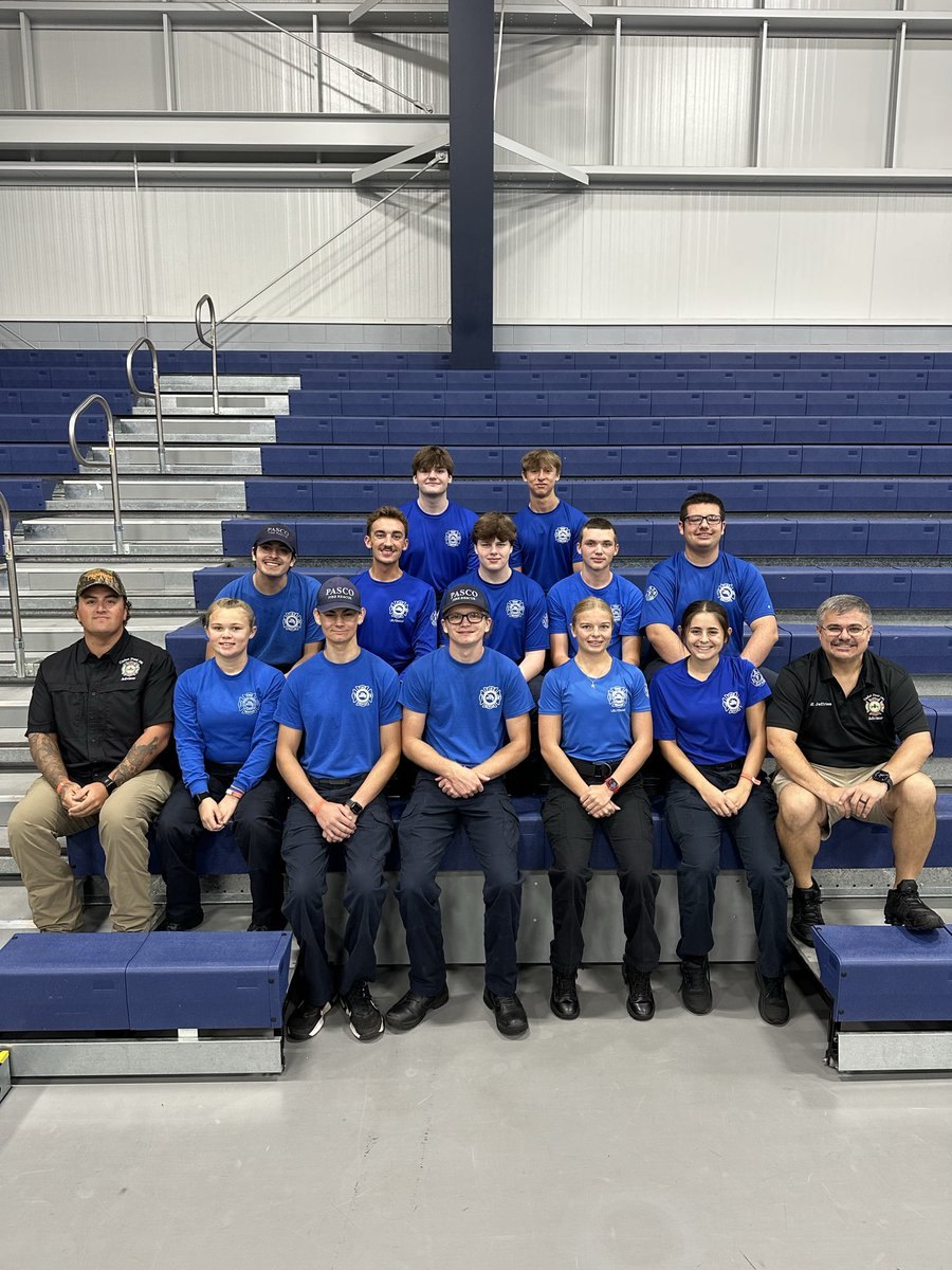 Group of teenagers in matching blue polo shirts posed on bleachers inside a sports facility.