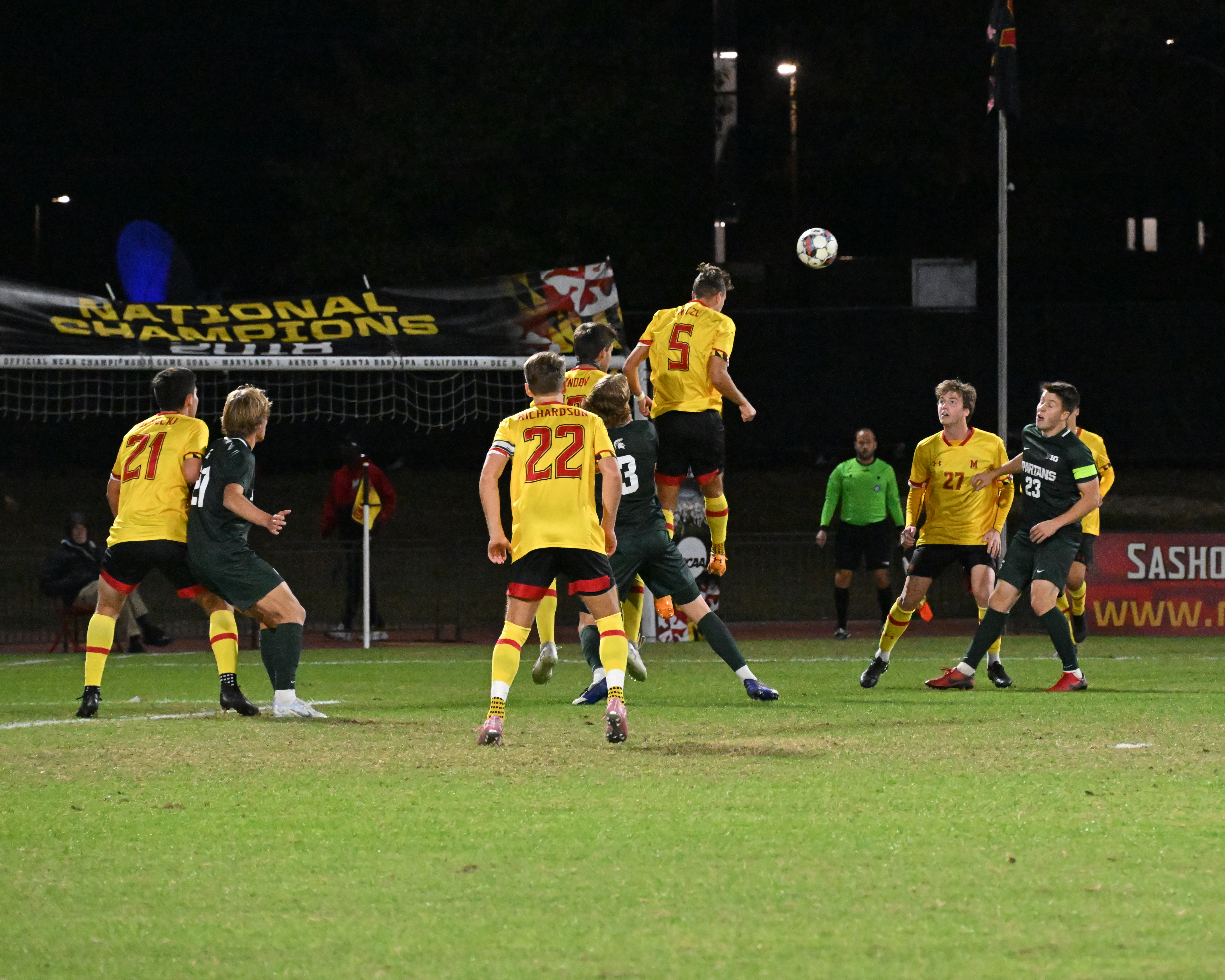 Two college soccer players leap for a header under stadium lights at night.