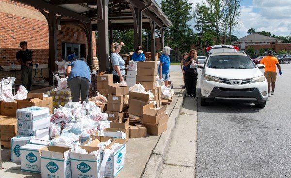 Volunteers passing cardboard boxes and bags to a white SUV at an outdoor distribution area.