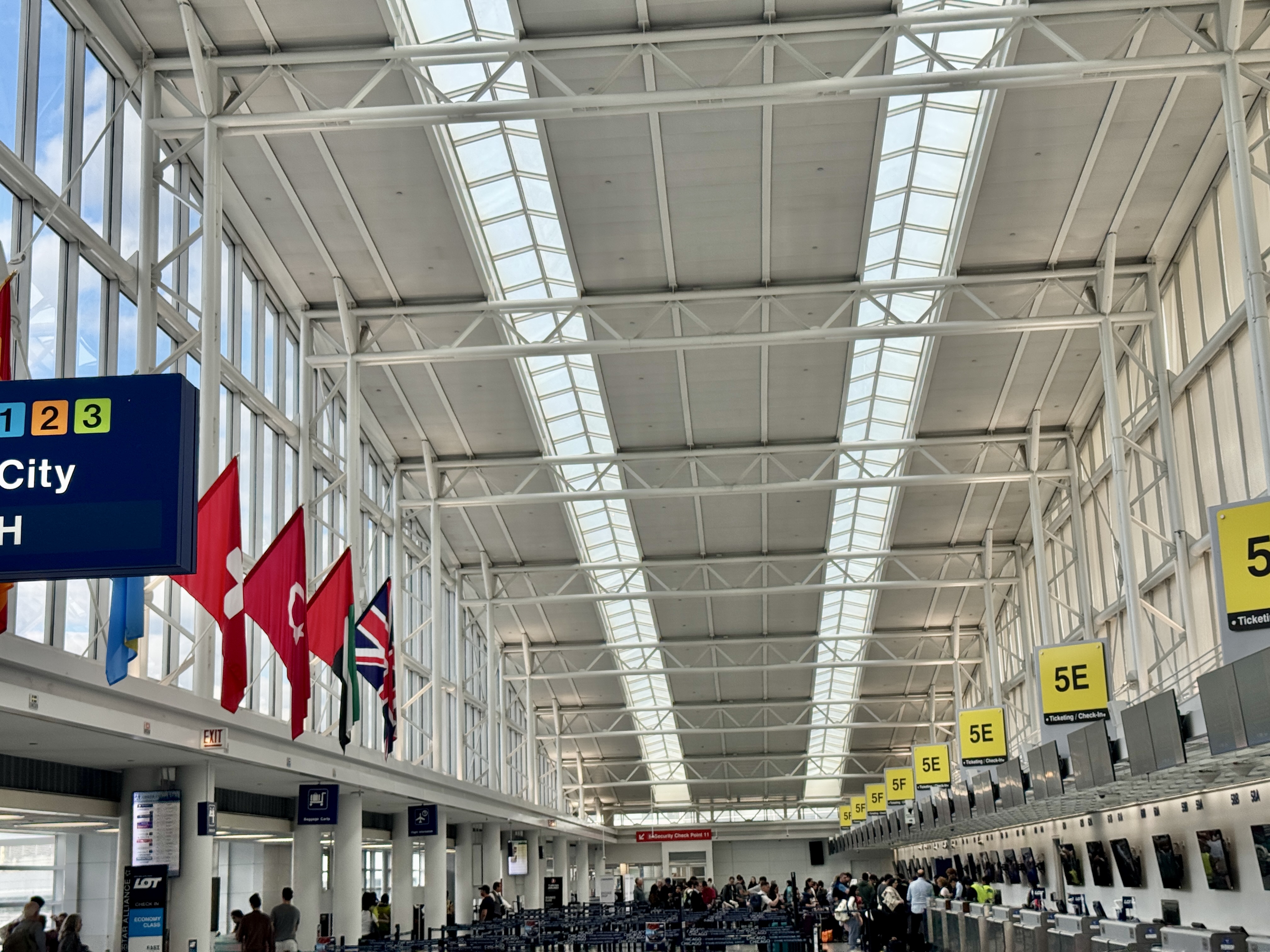 Airport terminal entrance hall with check-in counters, international flags hanging from ceiling, and travelers.