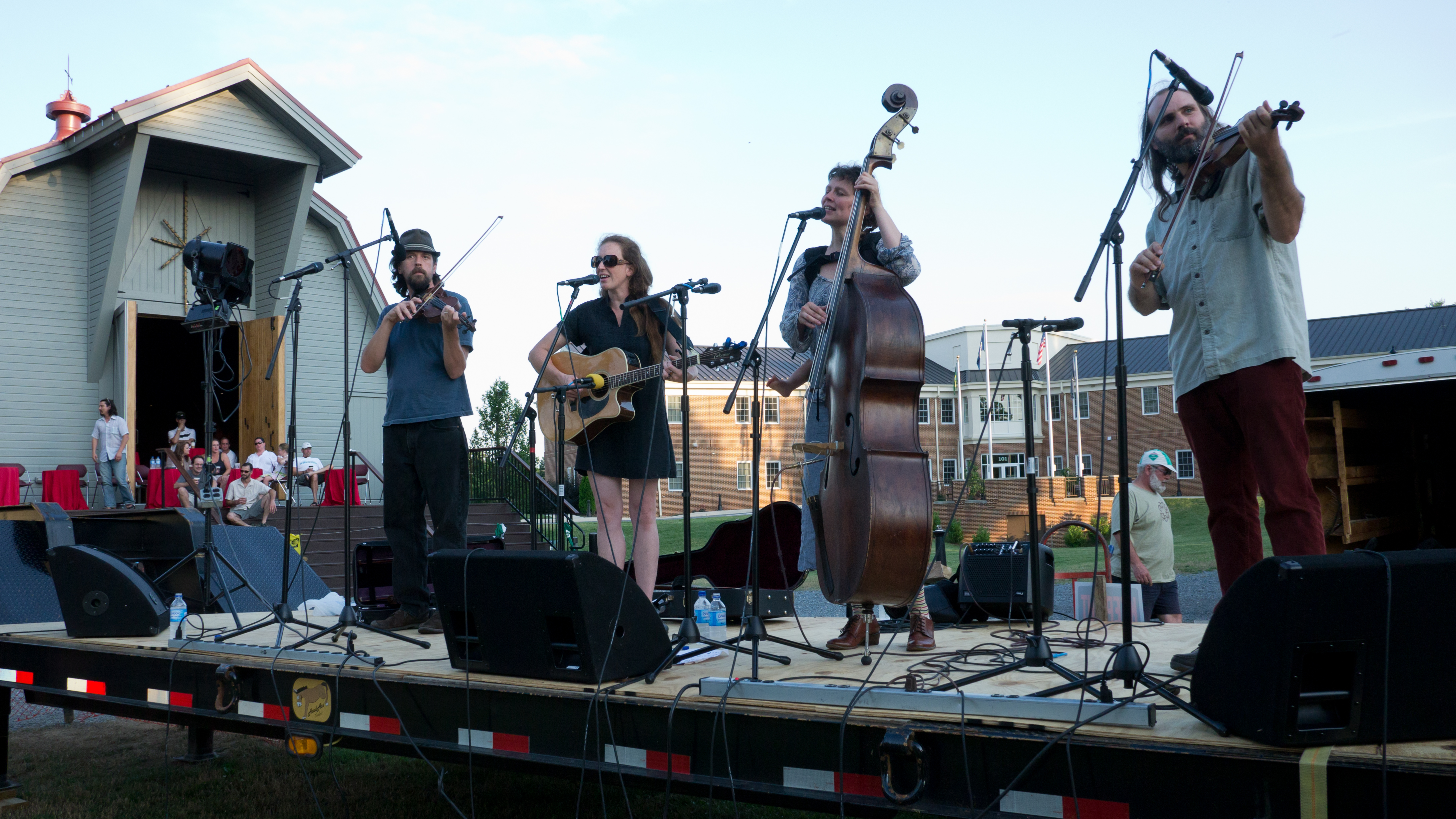 Band performing on an outdoor mobile stage at dusk with audience in background.