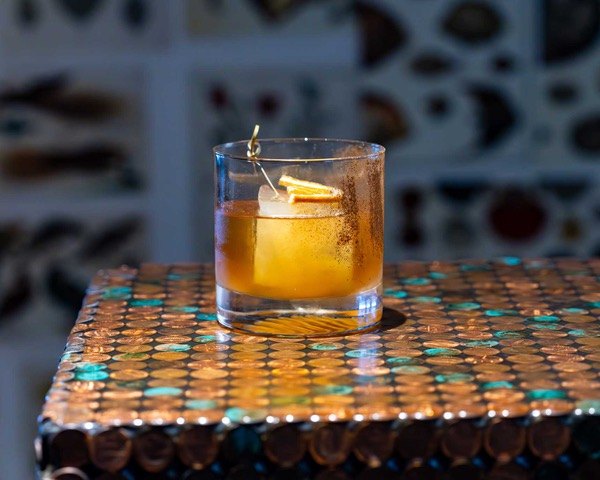 An amber cocktail in a rocks glass garnished with a citrus peel on a mosaic tile surface.