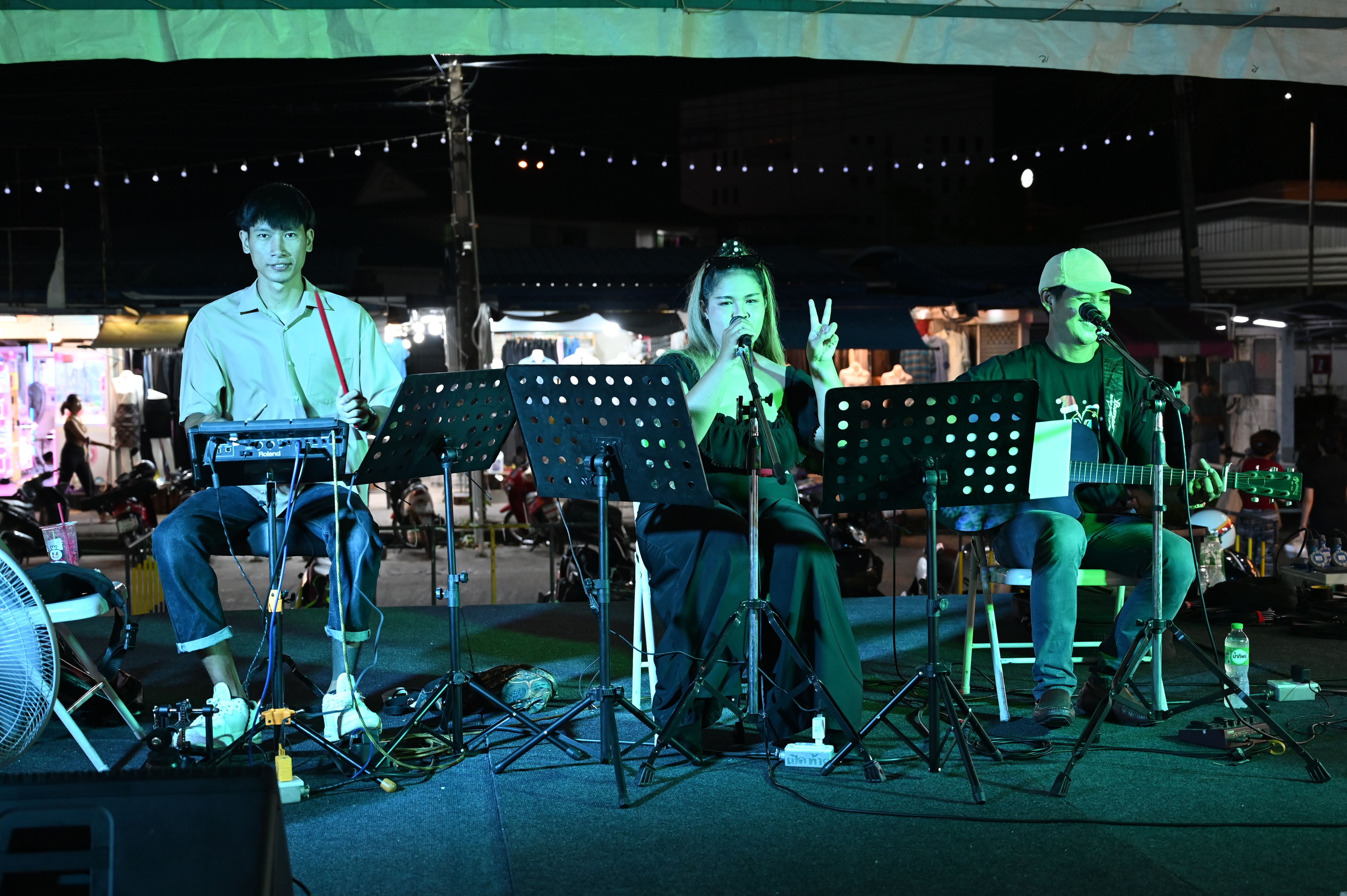 Three musicians perform on a small outdoor stage at night with string lights overhead.