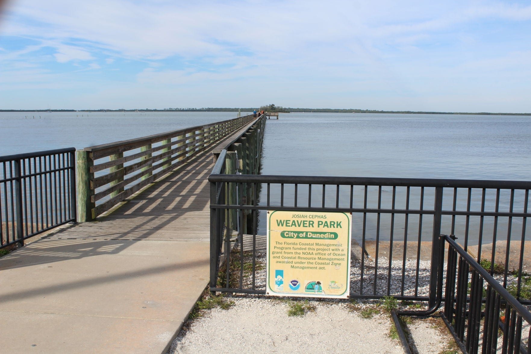A wooden pier extends over calm water at Weaver Park, with a park sign and metal railings visible along the sandy shoreline.