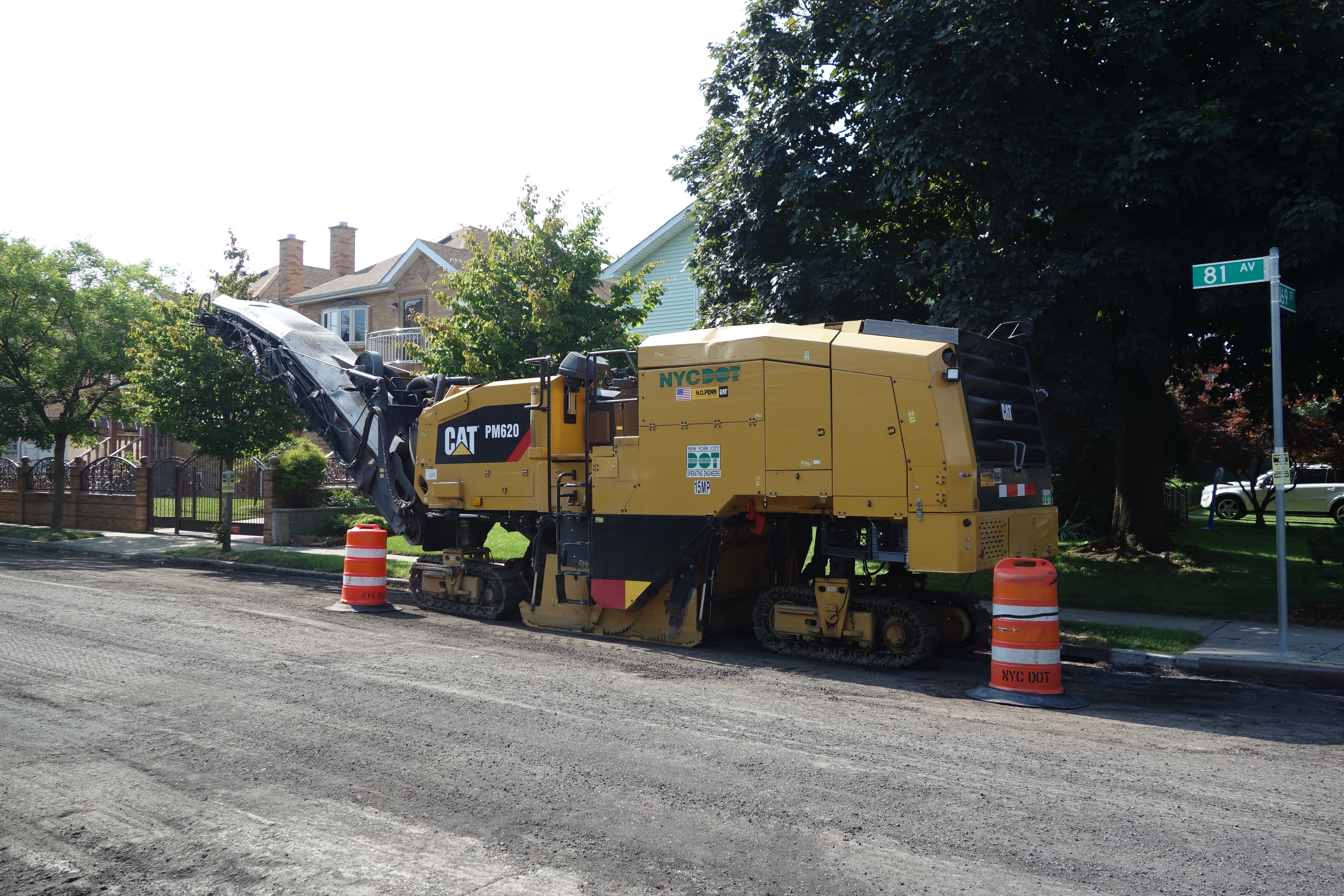 Yellow cold planer machine milling asphalt on a residential street with orange safety barrels.