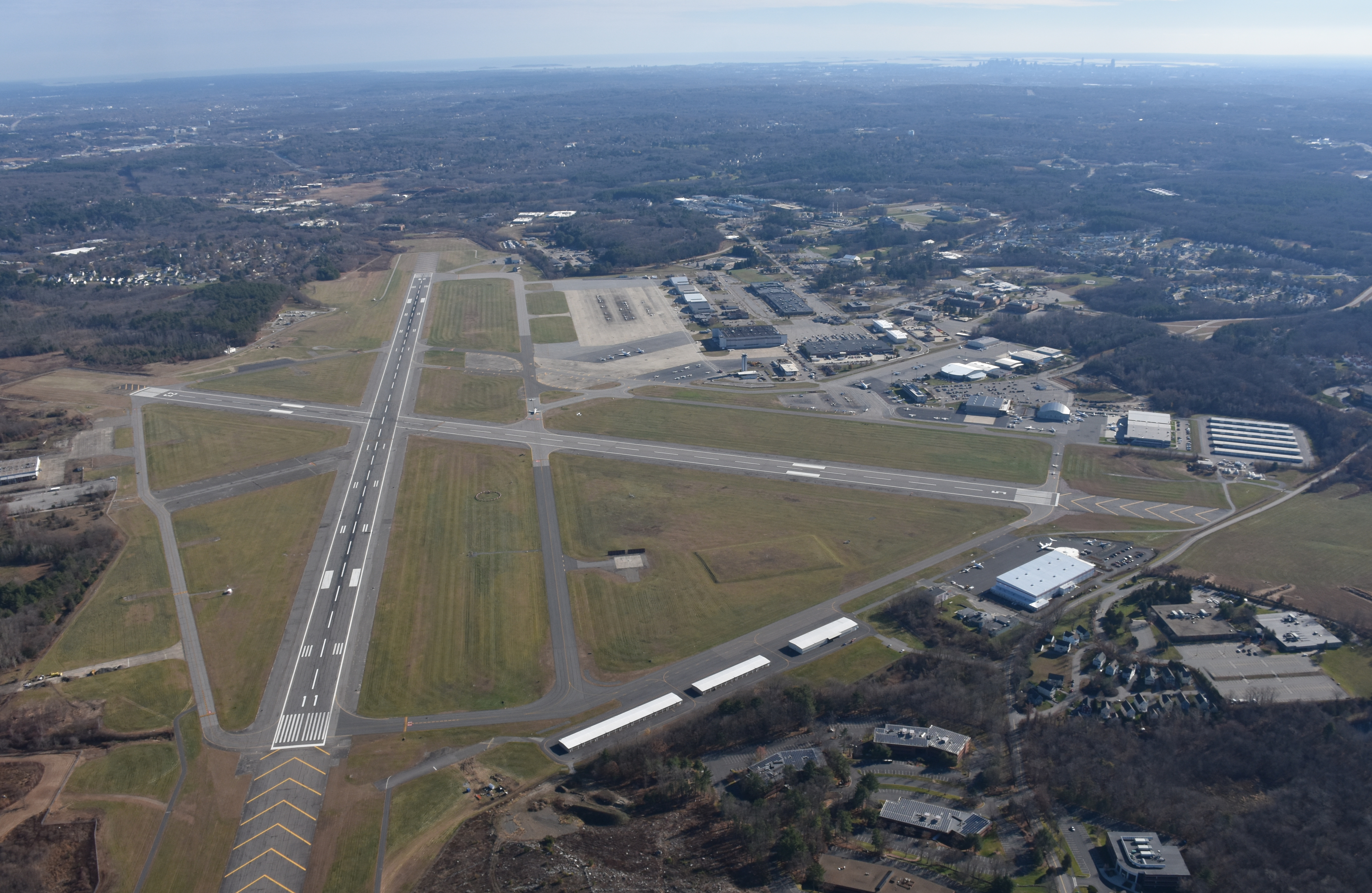 Aerial view of an airfield with two intersecting runways, hangars, and surrounding wooded suburbs.