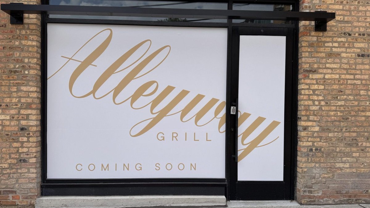 Storefront window on a brick building displaying a gold-script "Coming Soon" restaurant sign.