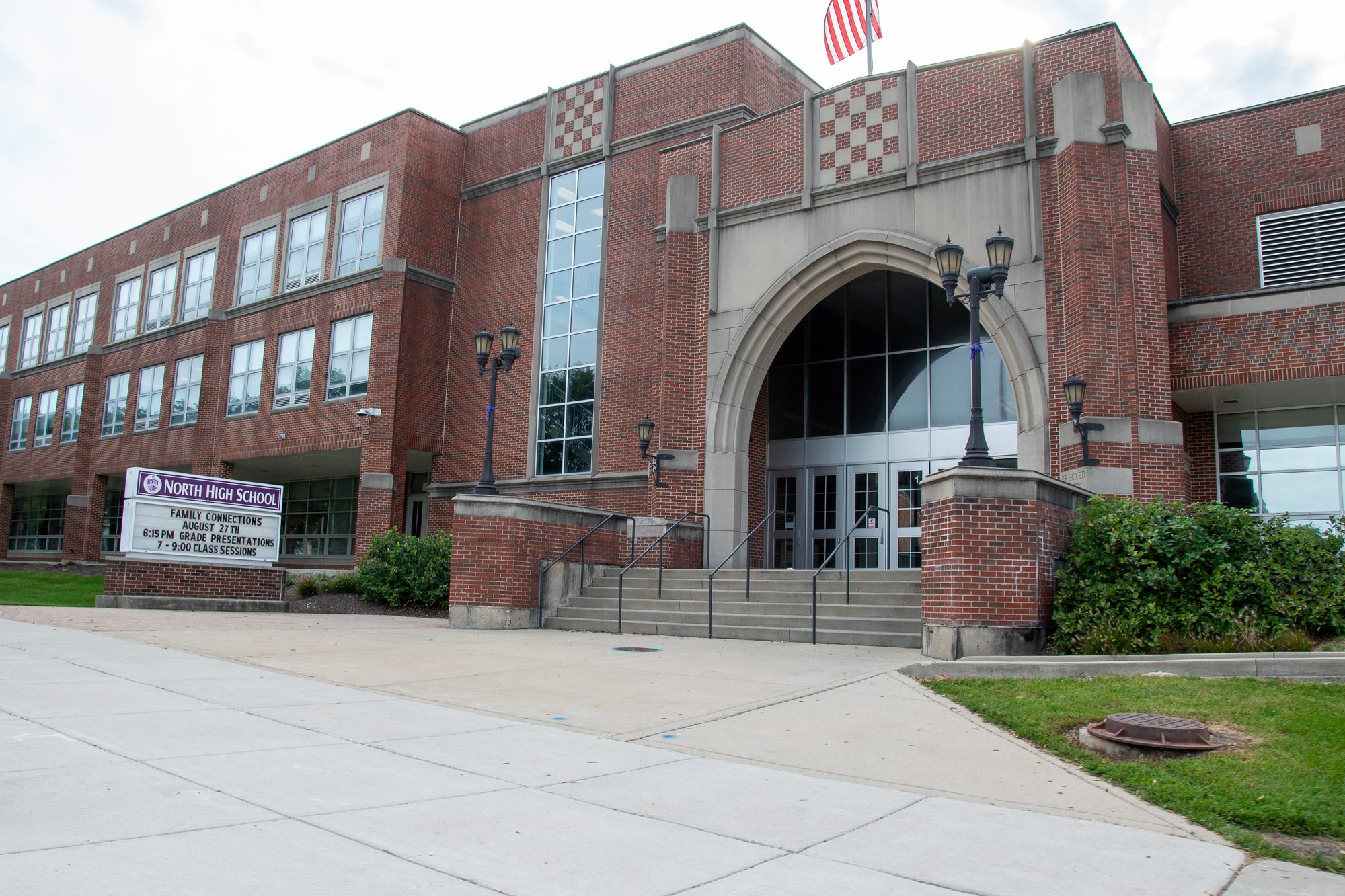 Exterior of a red-brick high school building with a large arched entrance and an American flag.