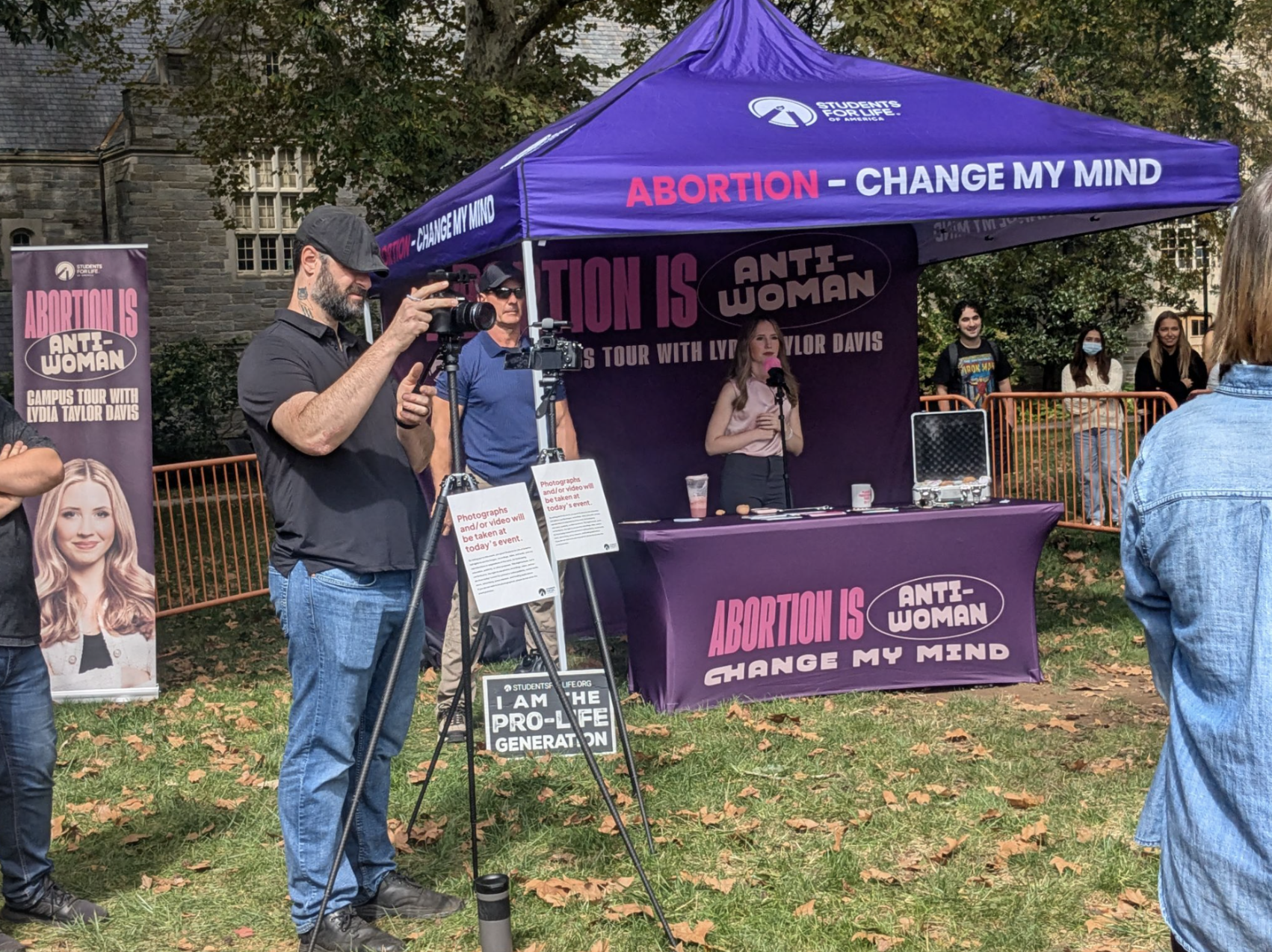 A purple tent with debate banners on a campus lawn, with a woman at a table and onlookers gathered around.
