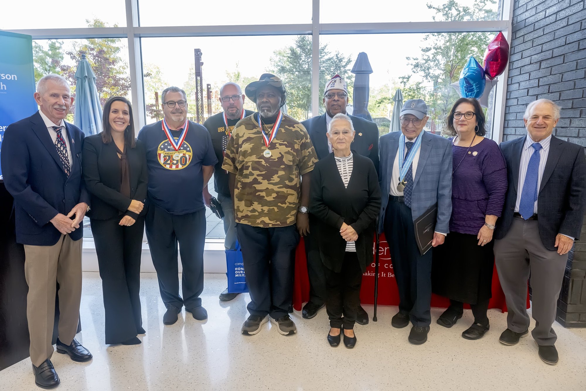 Group of adults posing indoors, several wearing large medals, with red, white, and blue balloons in the background.