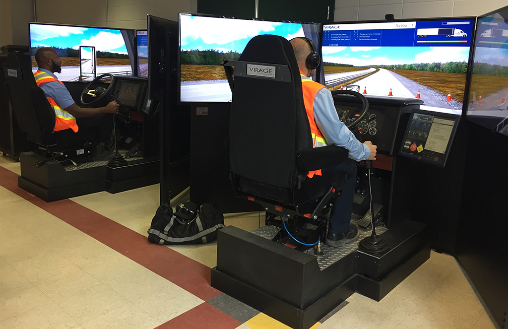 Two people in safety vests seated at multi-screen truck driving simulators indoors.