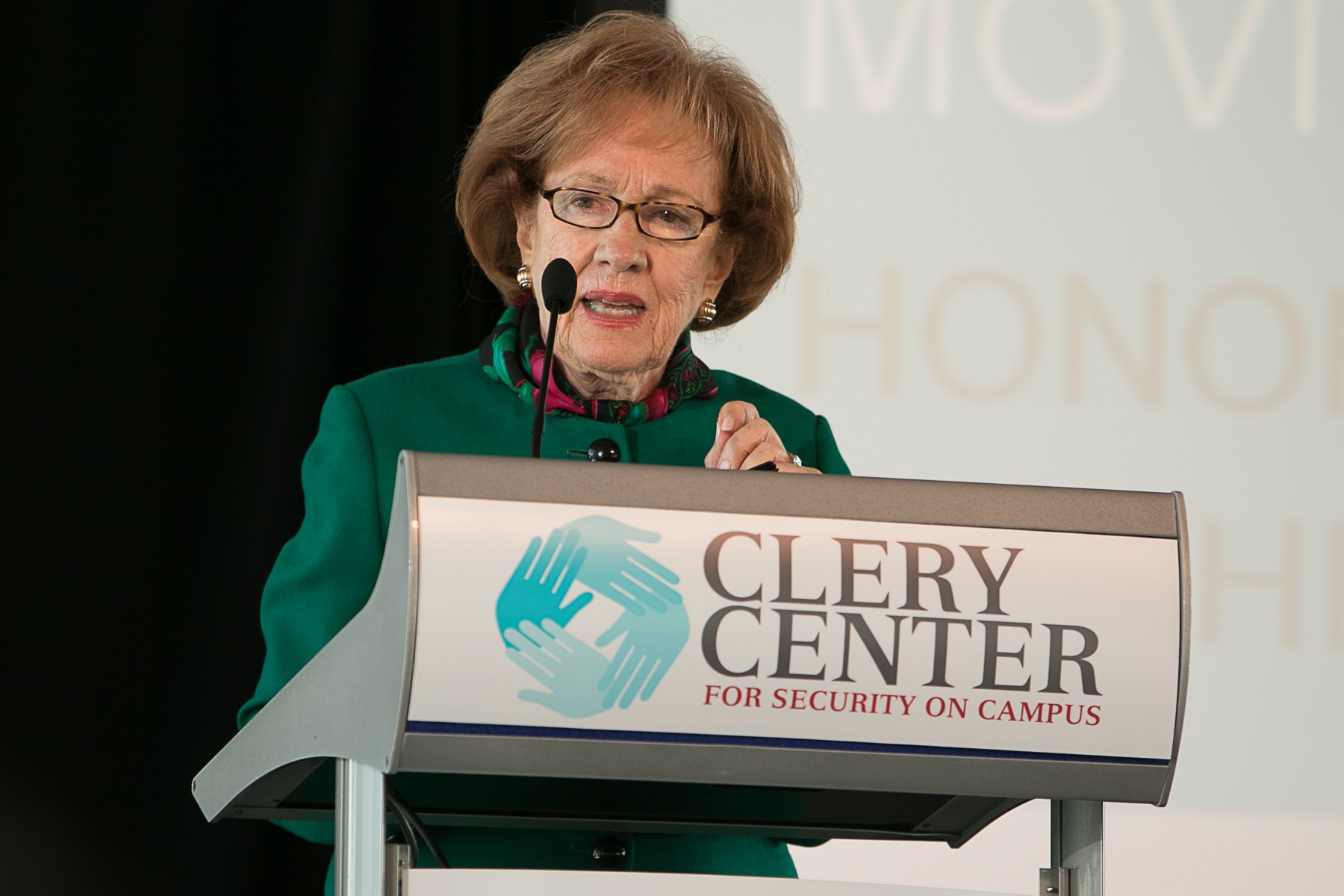 A woman in a green jacket speaks at a podium bearing the Clery Center for Security on Campus logo.
