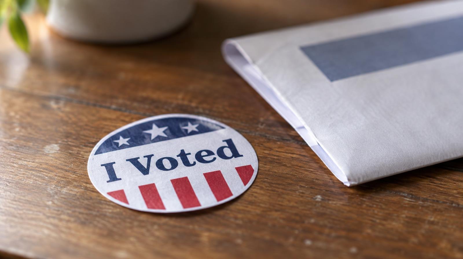 An I Voted sticker resting on a wooden surface beside a folded ballot envelope.