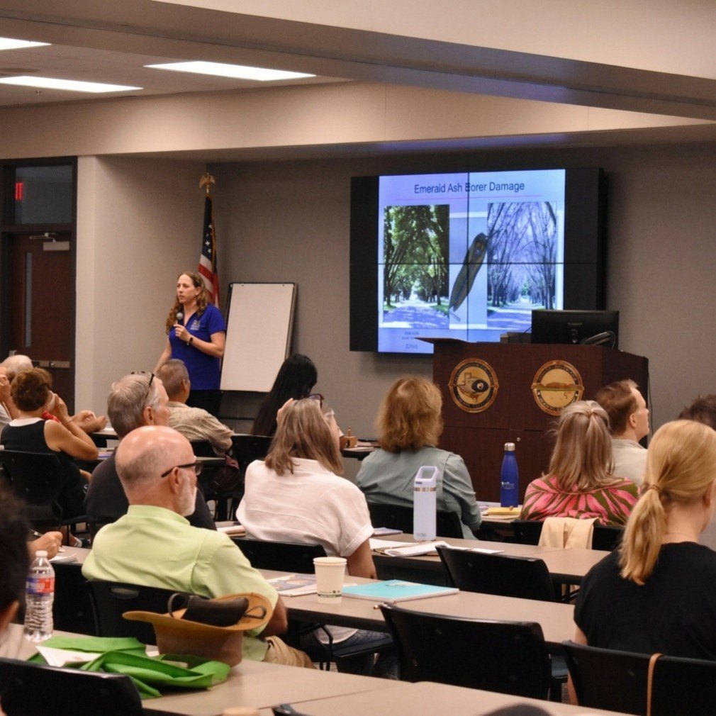 A woman presents an invasive species slideshow to a seated classroom audience.