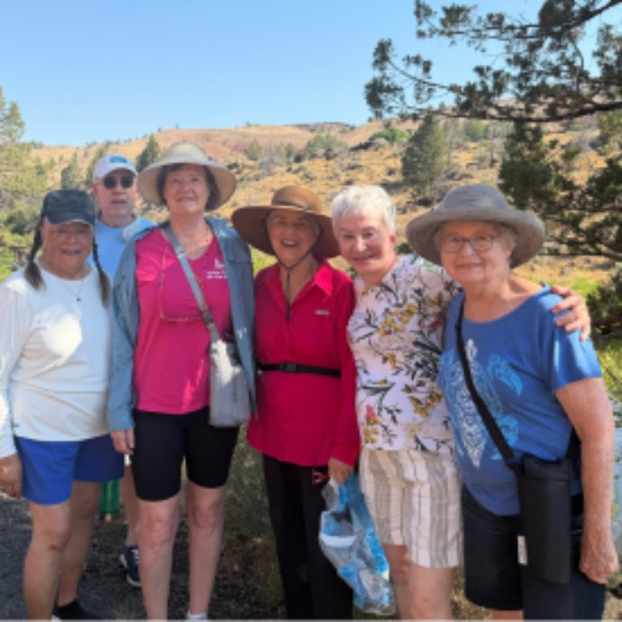 Six older adults pose together outdoors on a sunny day, wearing hats and casual clothing.