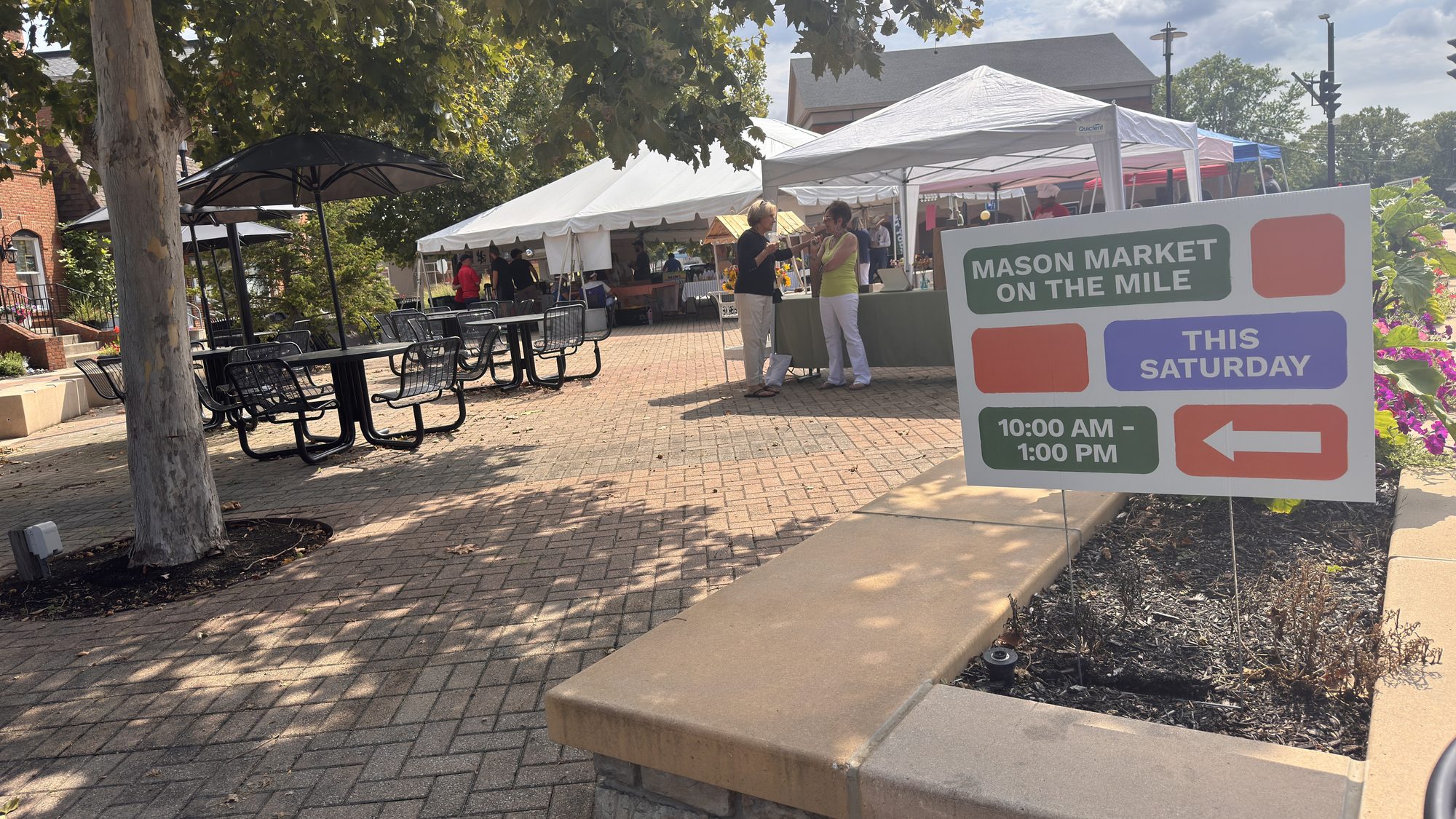 Outdoor market sign in a brick plaza with a white event tent and attendees in the background.