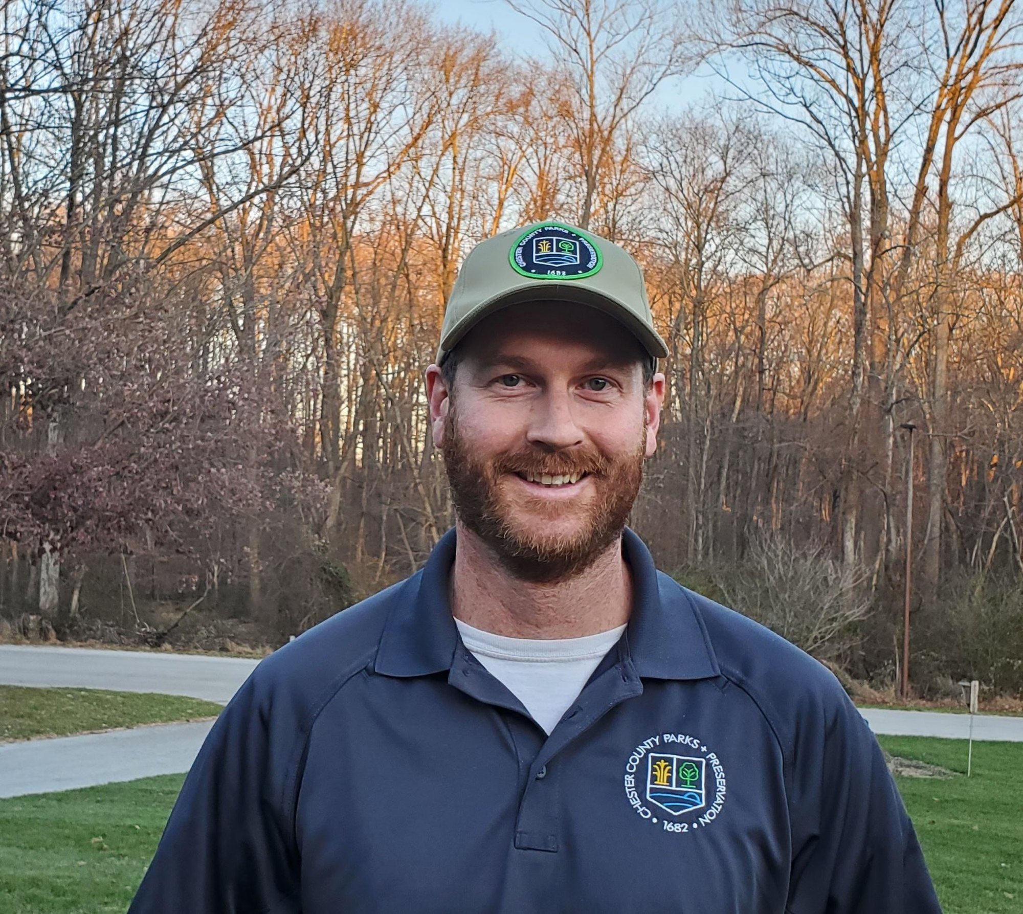 Smiling man in a navy Parks and Trails polo shirt and tan cap standing outdoors.